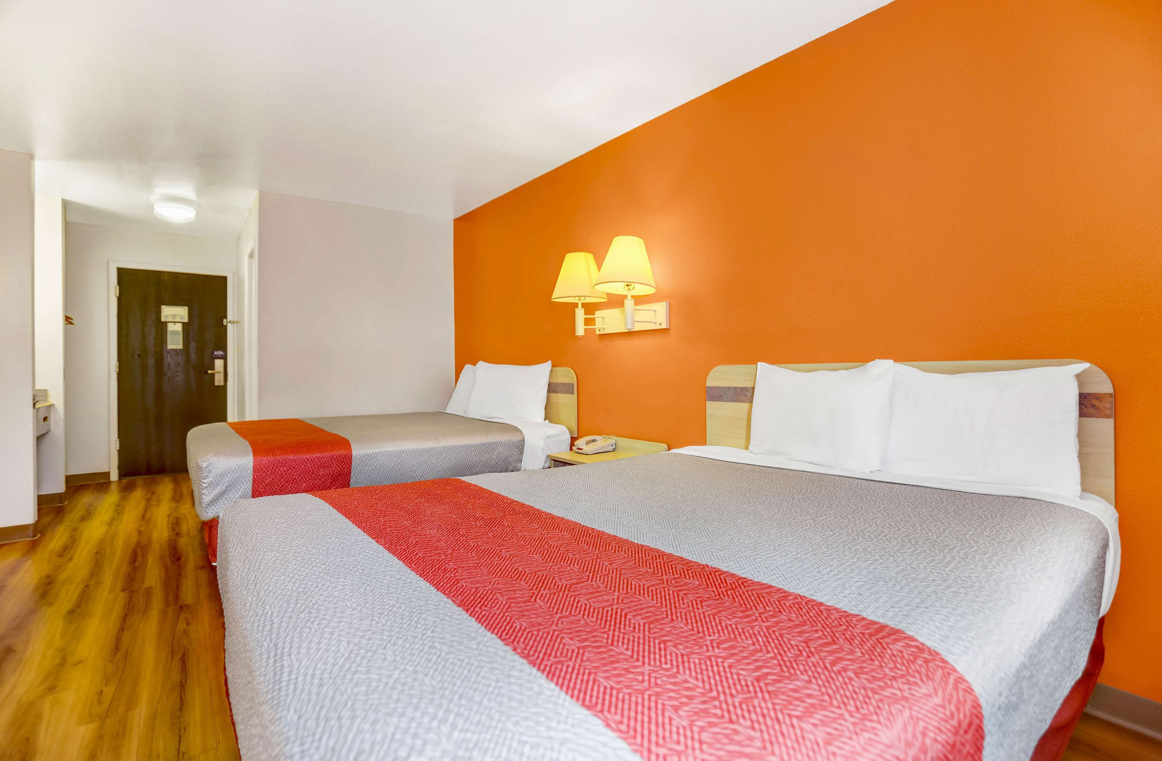 Een hotelkamer met twee bedden, een houten vloer, een oranje accentmuur en een nachtkastje met een lamp.