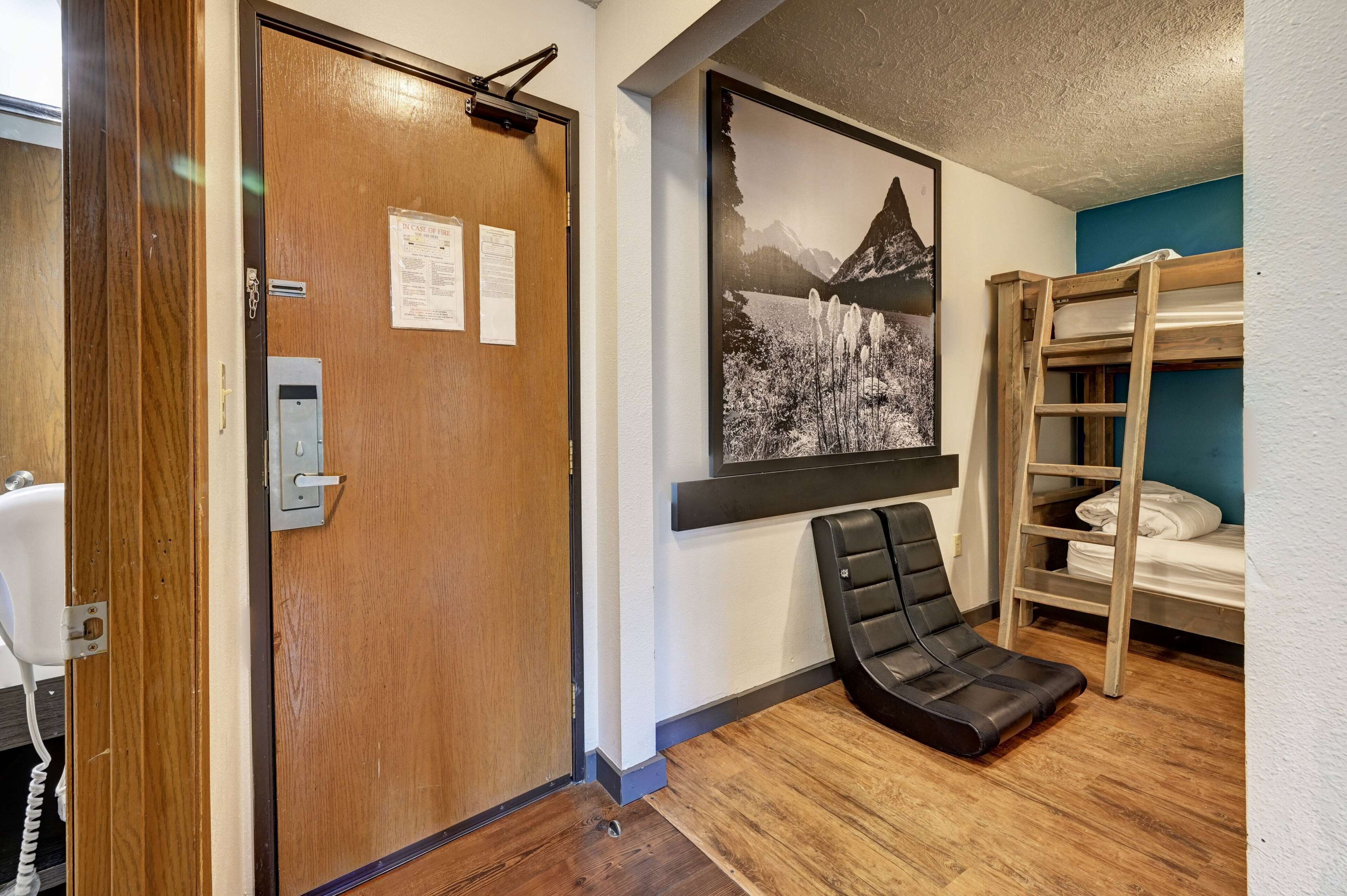 Habitación de hotel con puerta de madera, un cuadro colgado de un paisaje de montaña, una cama litera y una butaca negra.