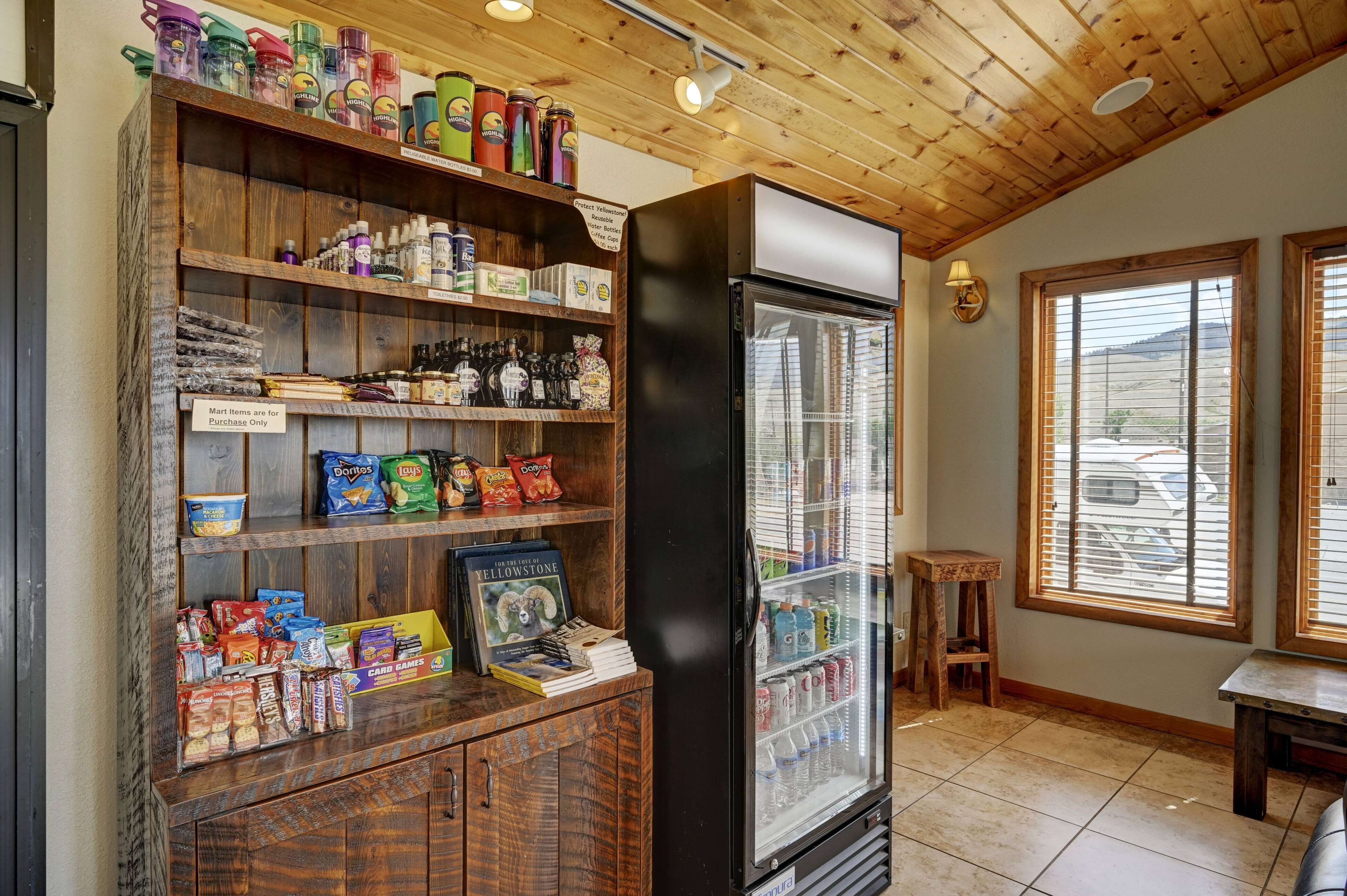 A store with a wooden interior, shelves stocked with snacks, a refrigerator, and a window with blinds.