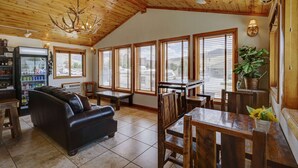 A spacious room with a high ceiling, wooden beams, a leather sofa, a dining table with chairs, and large windows with blinds.
