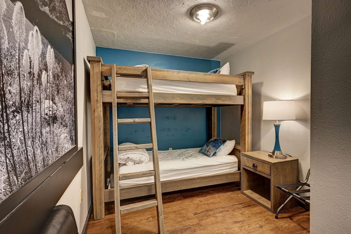 Una cama litera con escalera de madera, una mesita de noche con lámpara y una pared con un cuadro abstracto de gran tamaño.