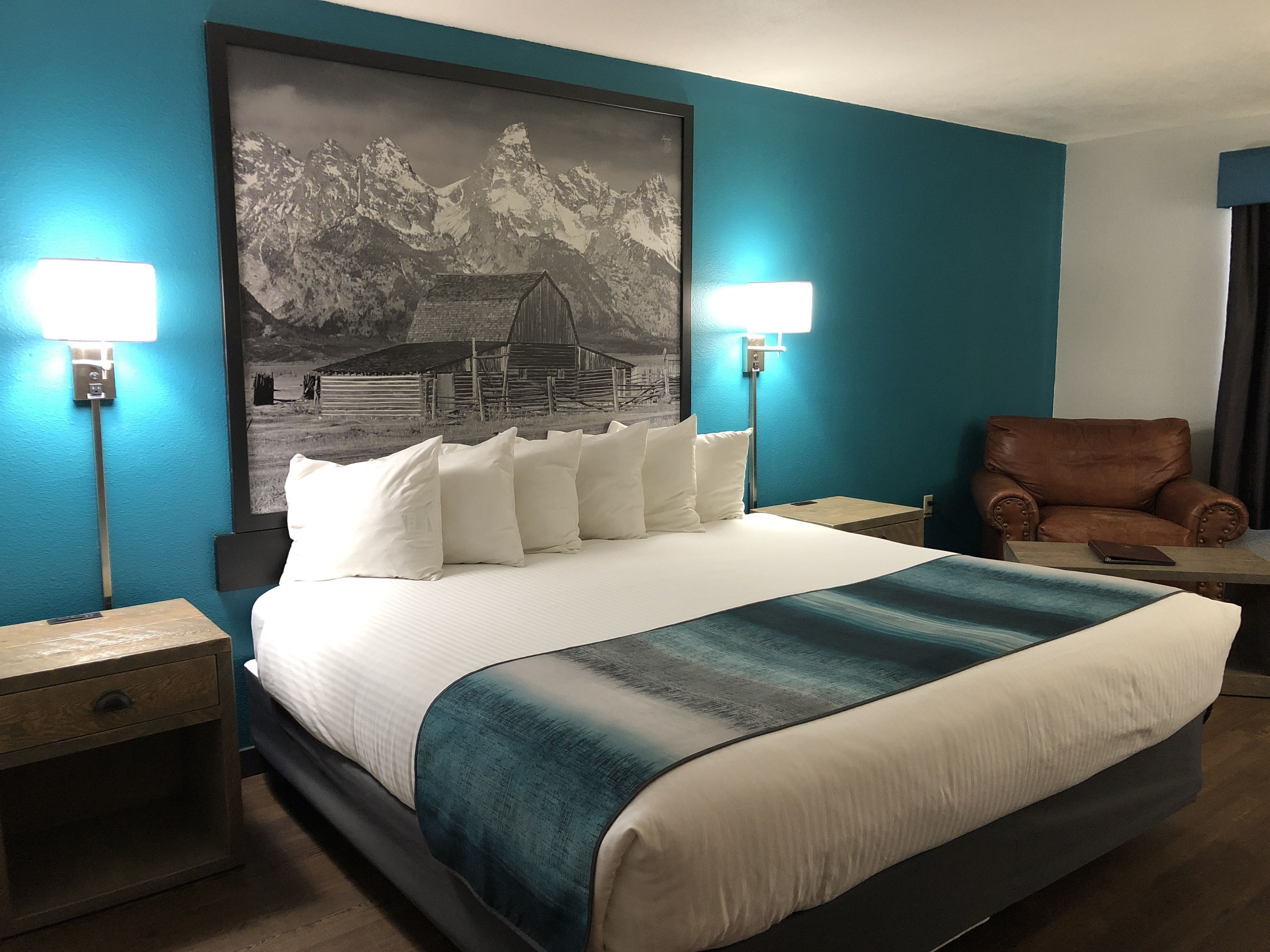 Een hotelkamer met een groot bed, twee bedlampen, een bruine leren fauteuil en een berglandschap op de muur.