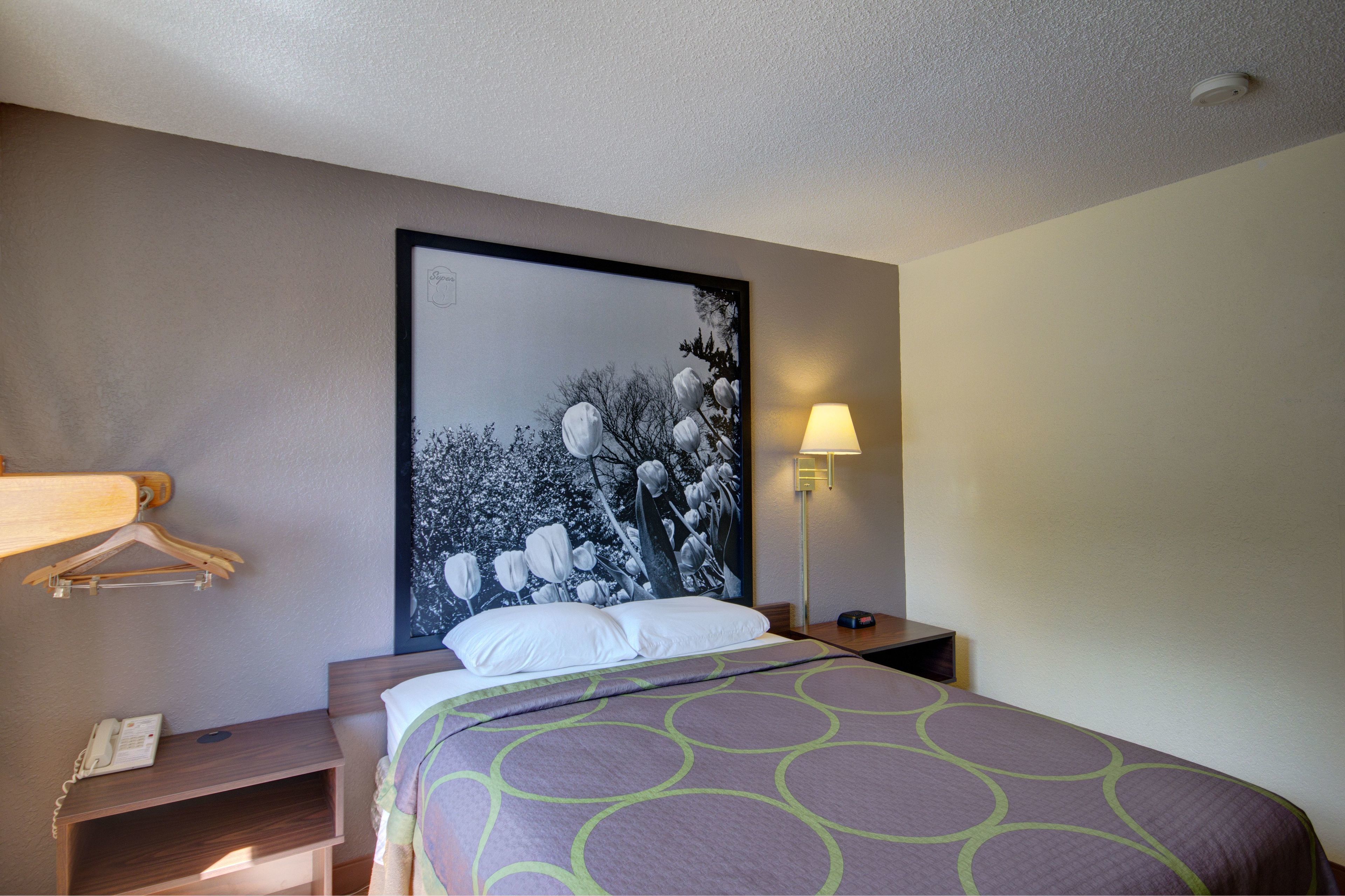 A hotel room with a bed, a nightstand, a lamp, a phone, and a wall-mounted coat rack.