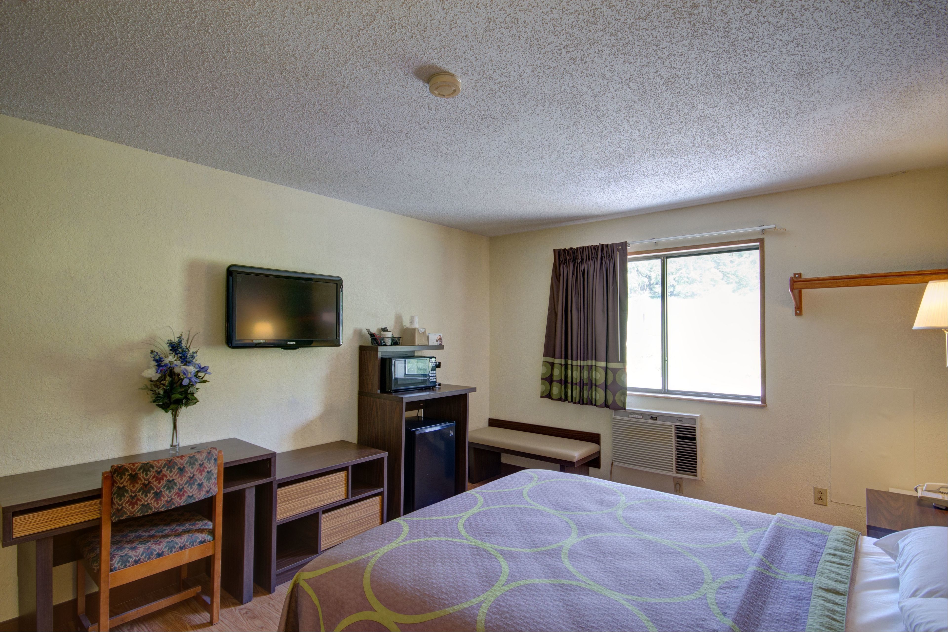 Una habitación de hotel con una cama, un escritorio con silla, un televisor, una ventana con cortinas y un aire acondicionado instalado en la pared.