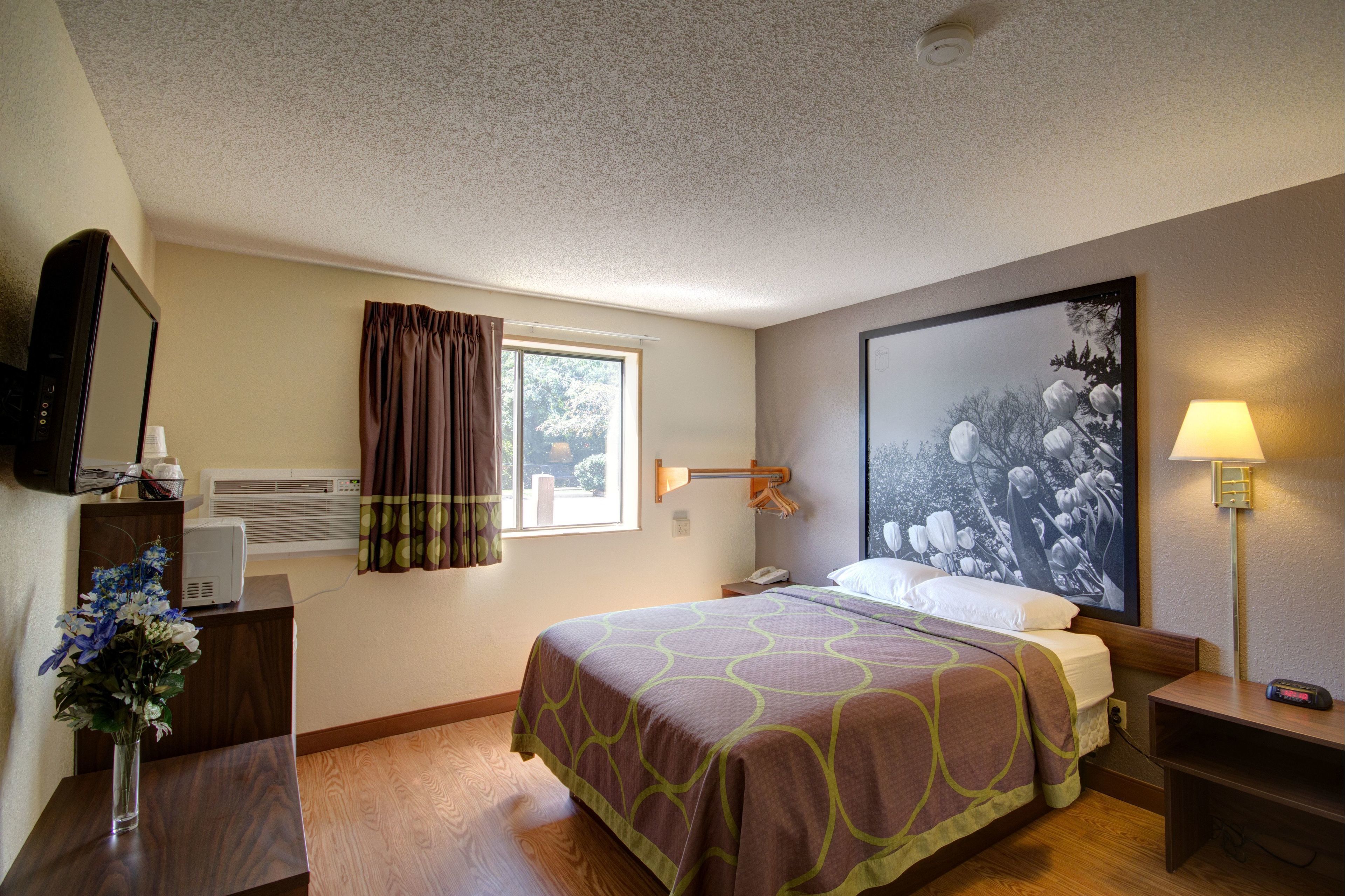 Una habitación de hotel con cama, escritorio con lámpara, ventana con cortinas, aire acondicionado de pared y un cuadro.