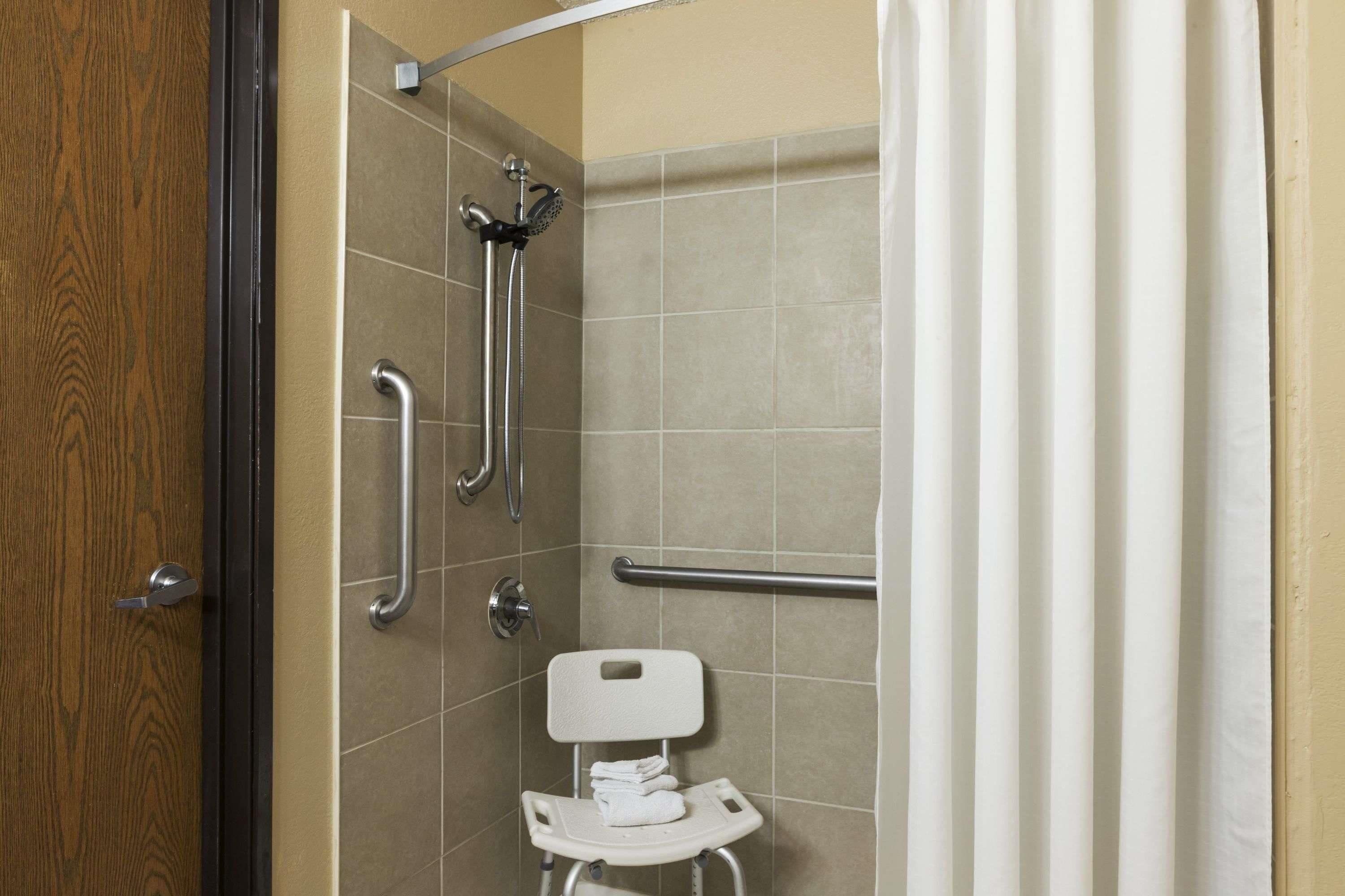 A bathroom with a shower, grab bars, and a white chair.