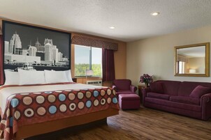Habitación de hotel con una cama grande, un cartel con un paisaje urbano, una ventana con cortinas, un sillón morado y un espejo en la pared.