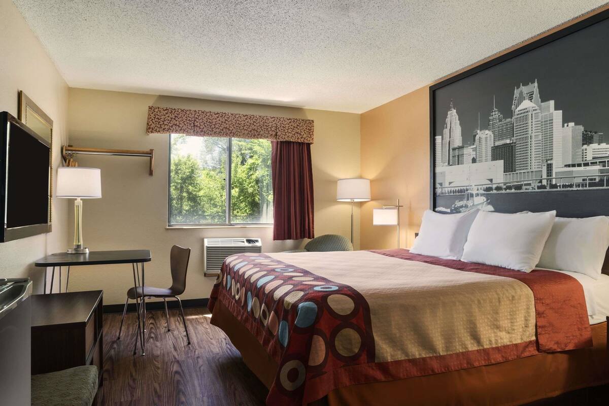 A hotel room with a bed, a desk, a chair, a window with a view of a city skyline, and a cityscape painting on the wall.