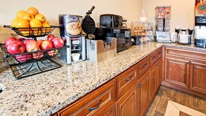 A kitchen with a fruit basket, waffle maker, coffee maker, and a refrigerator.