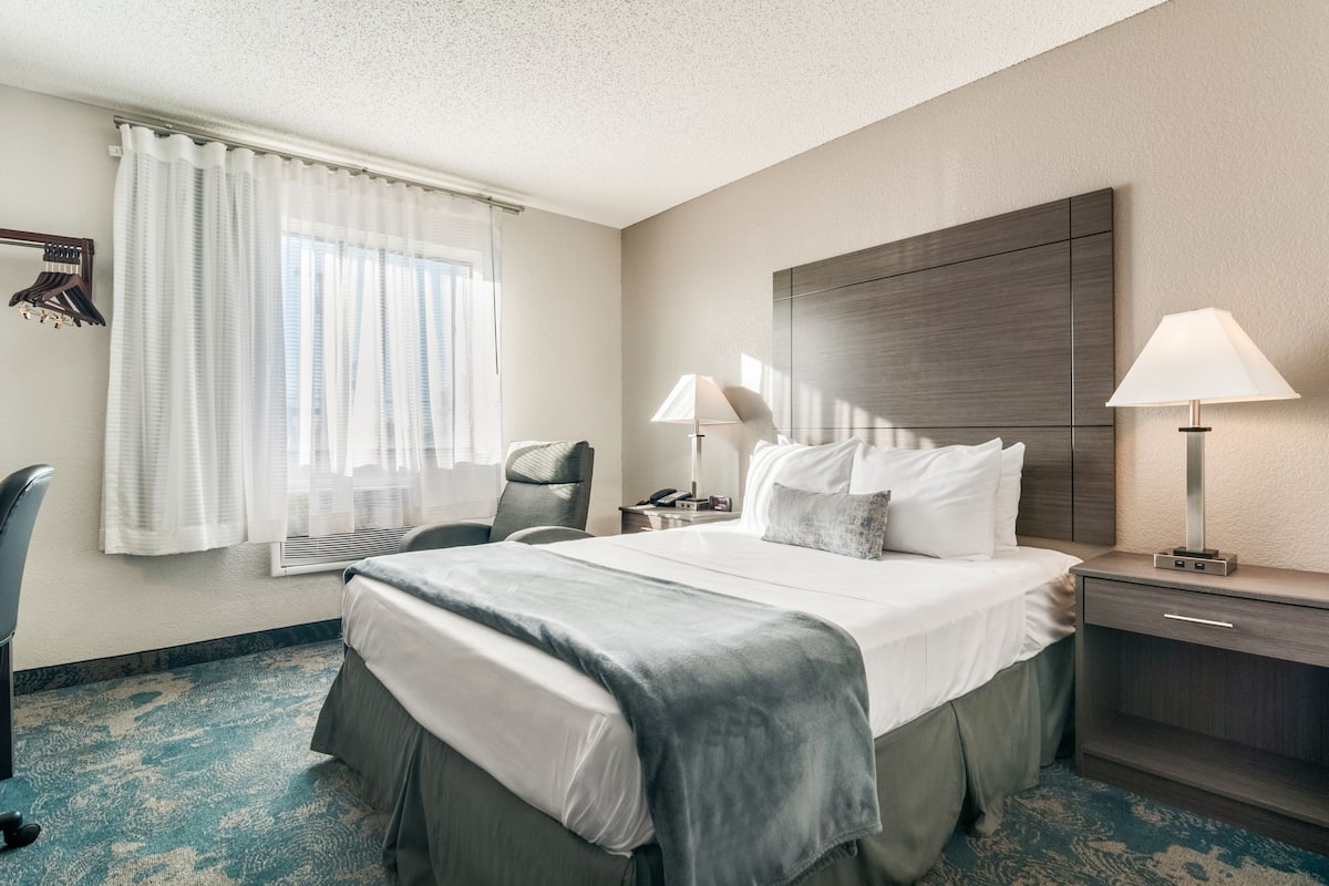 A hotel room with a large bed, two bedside tables with lamps, a desk, a chair, and a window with curtains.
