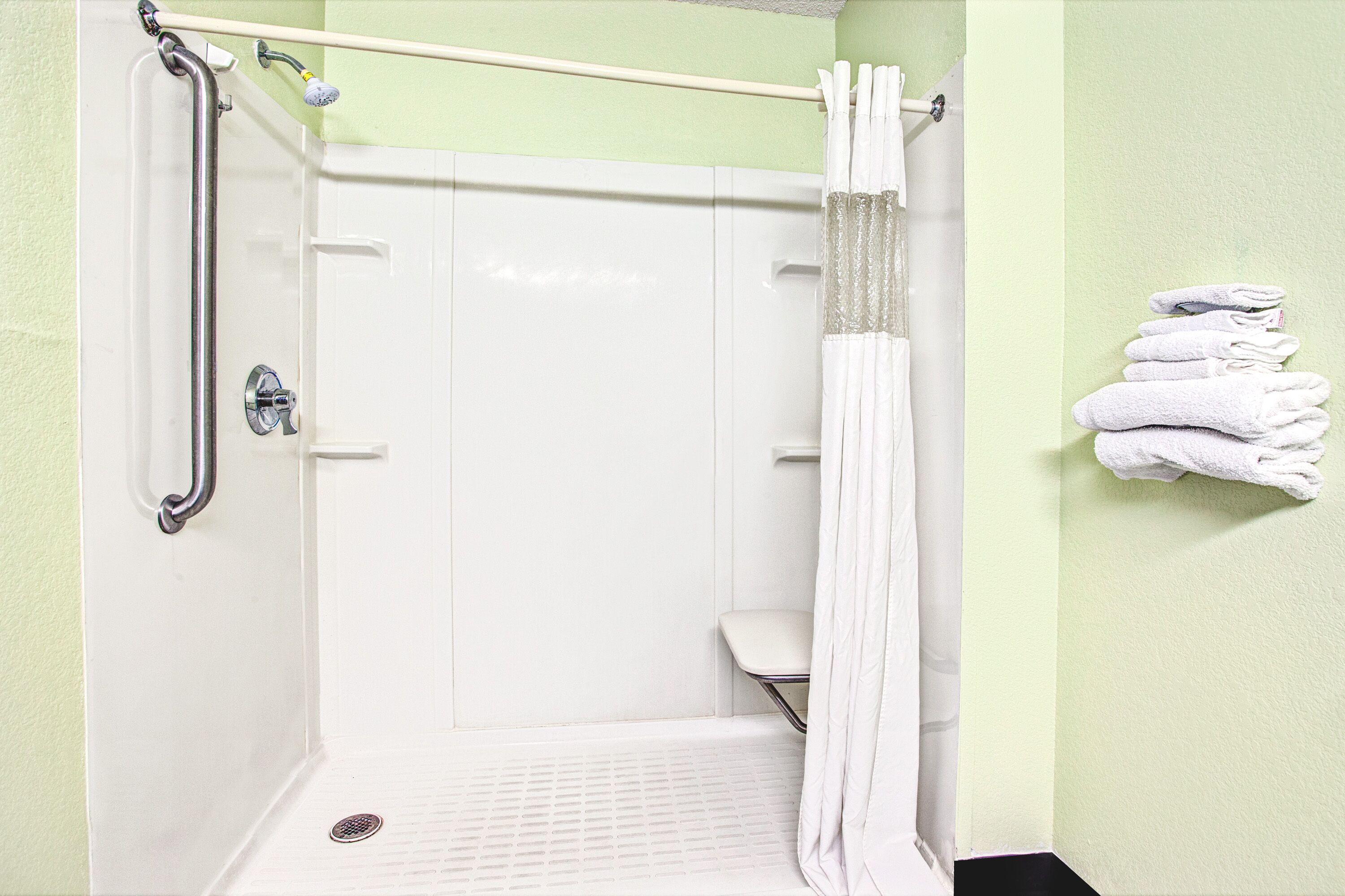 A shower area with a grab bar, a white shower curtain, and a stack of white towels on the wall.
