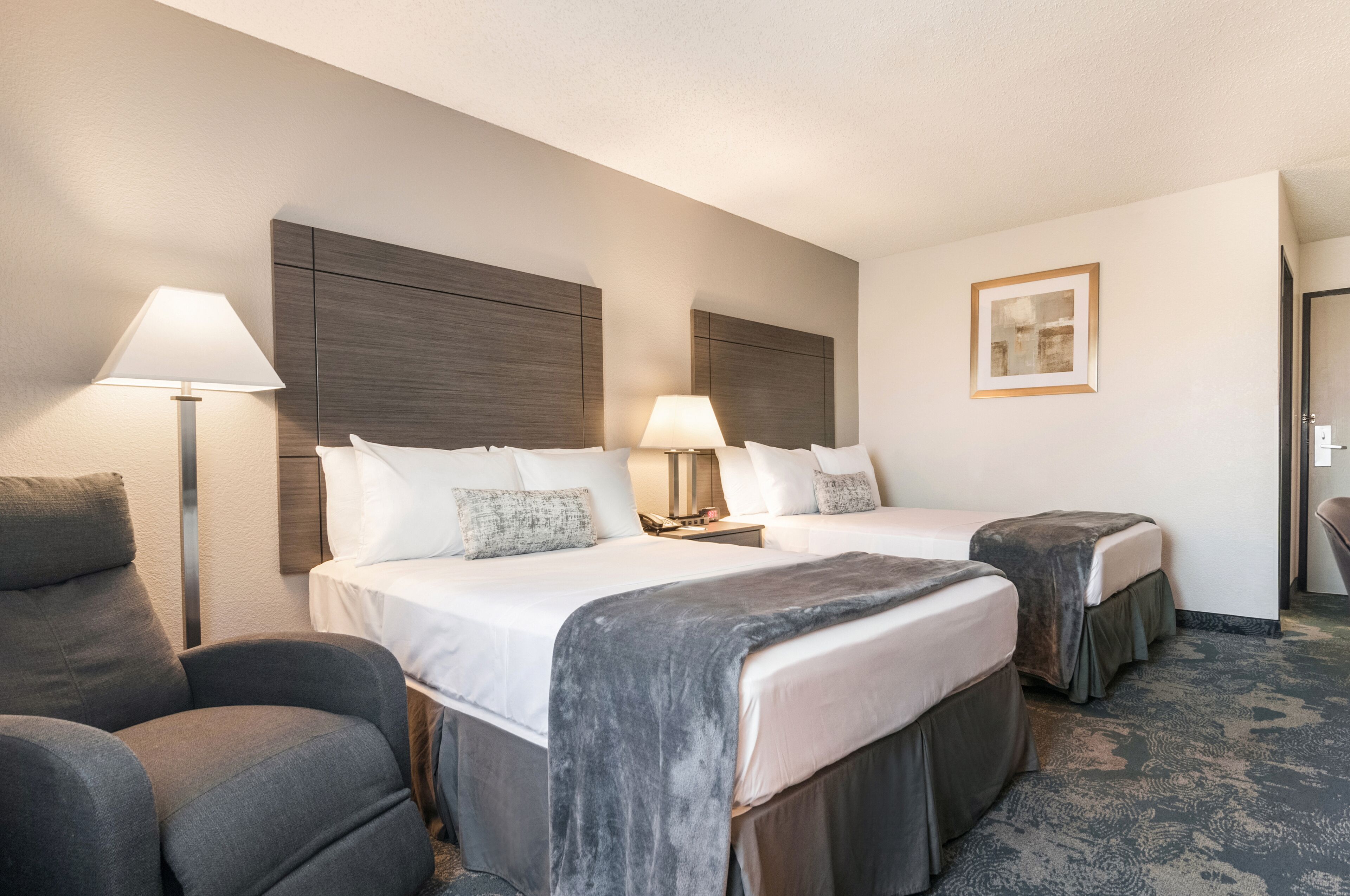 A hotel room with two beds, a desk, a chair, and a lamp.