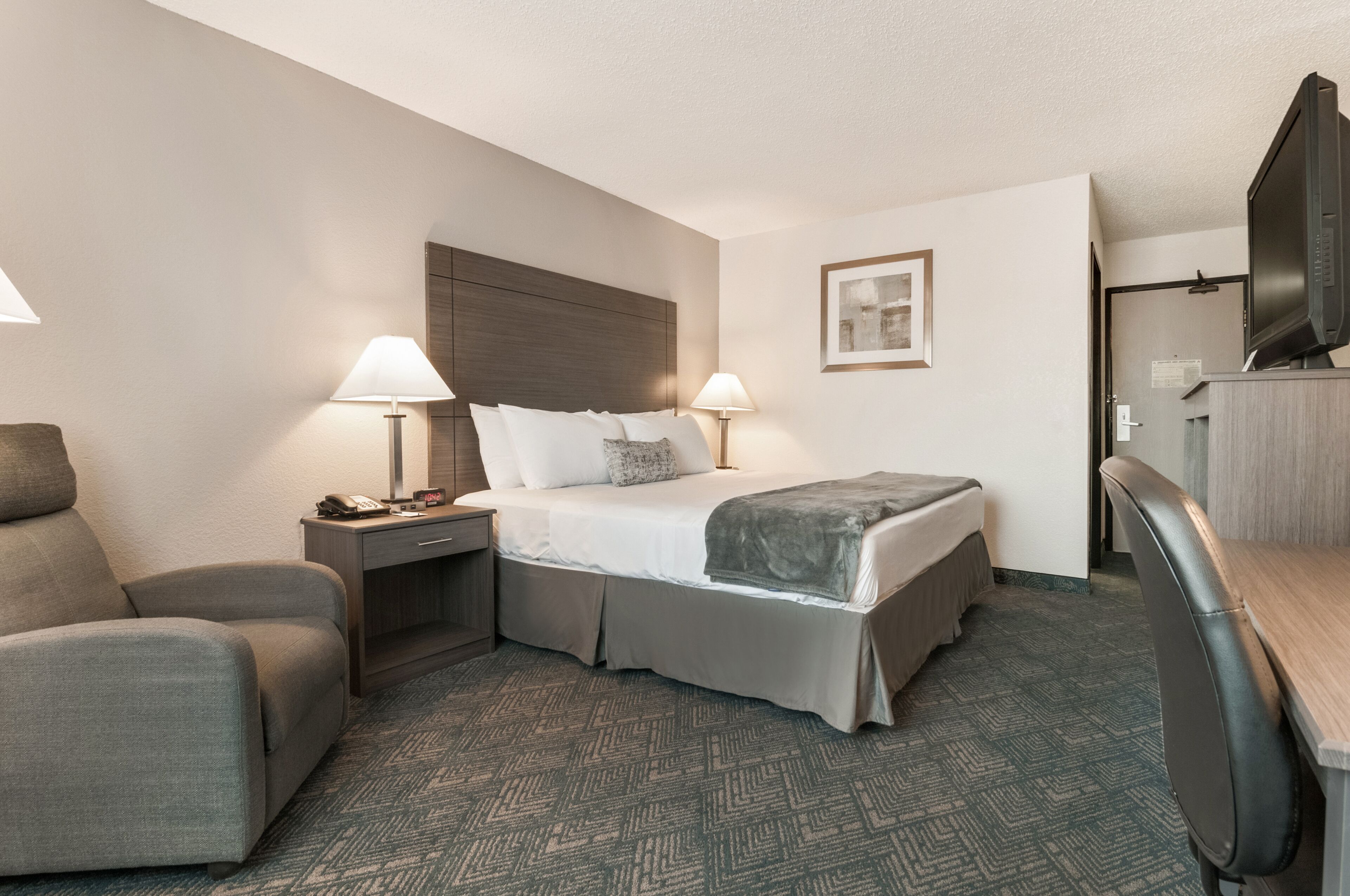 A hotel room with a bed, two bedside lamps, a desk with a chair, a TV, and a framed picture on the wall.