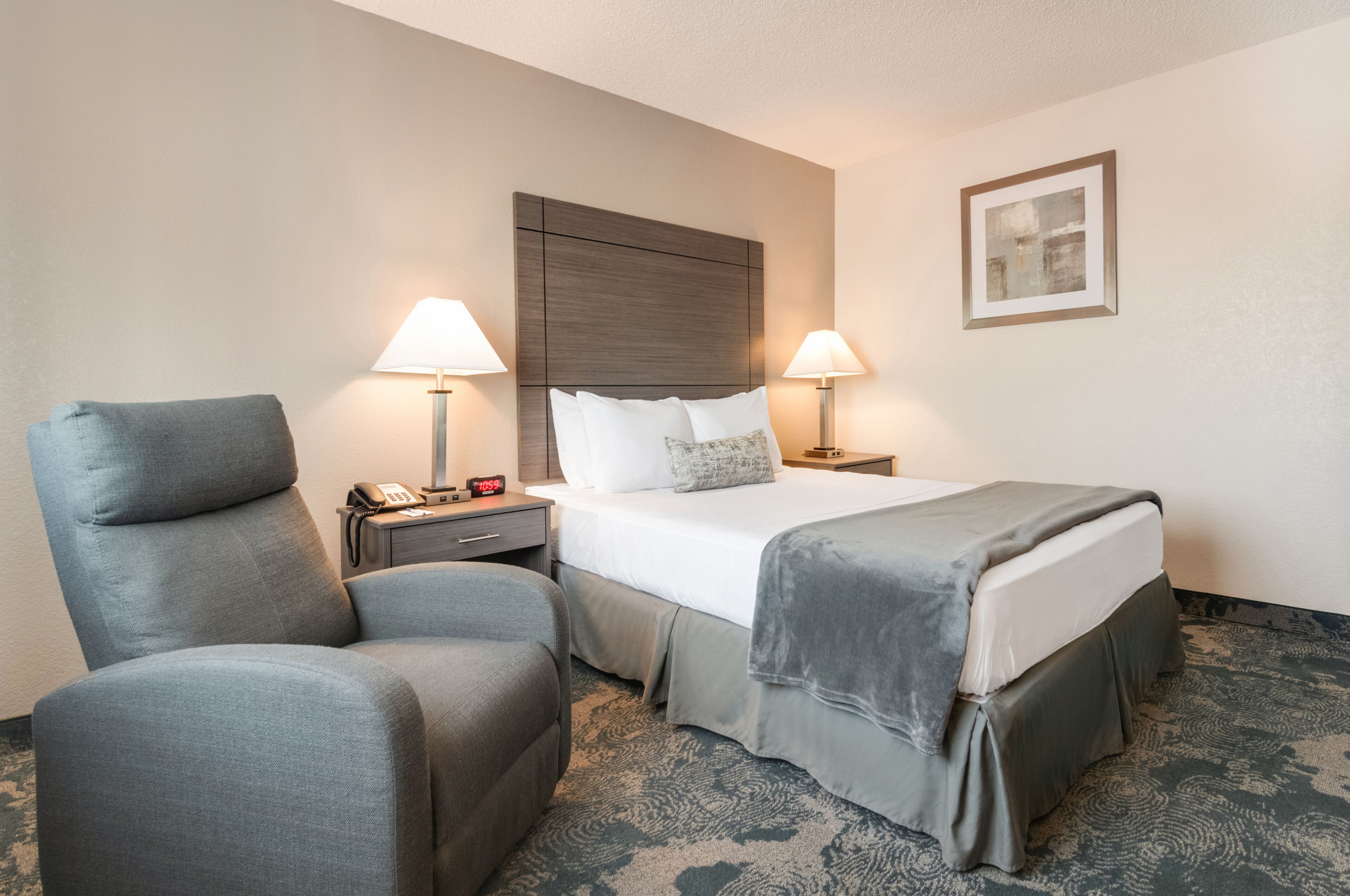 A hotel room with a bed, two bedside lamps, a nightstand with a phone, a chair, and a carpeted floor.