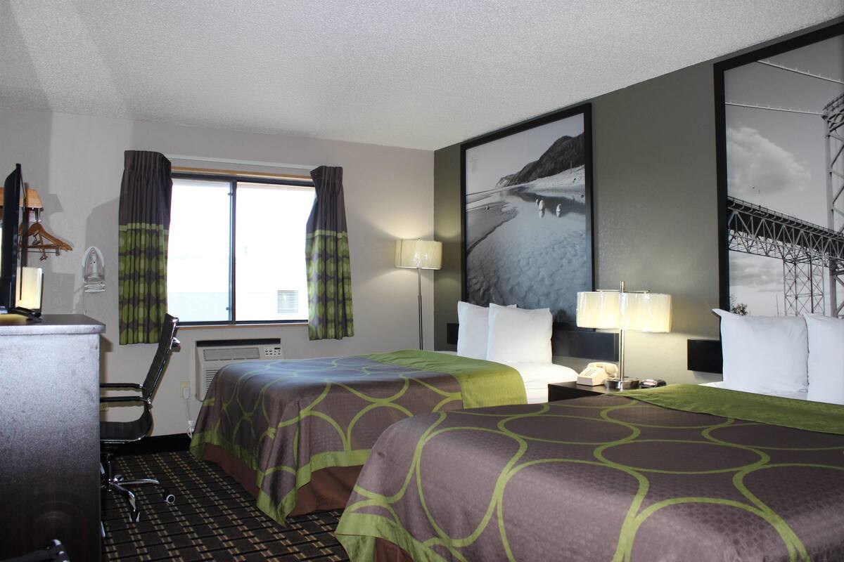 A hotel room with two beds, a desk, a chair, a lamp, and a large window with curtains.