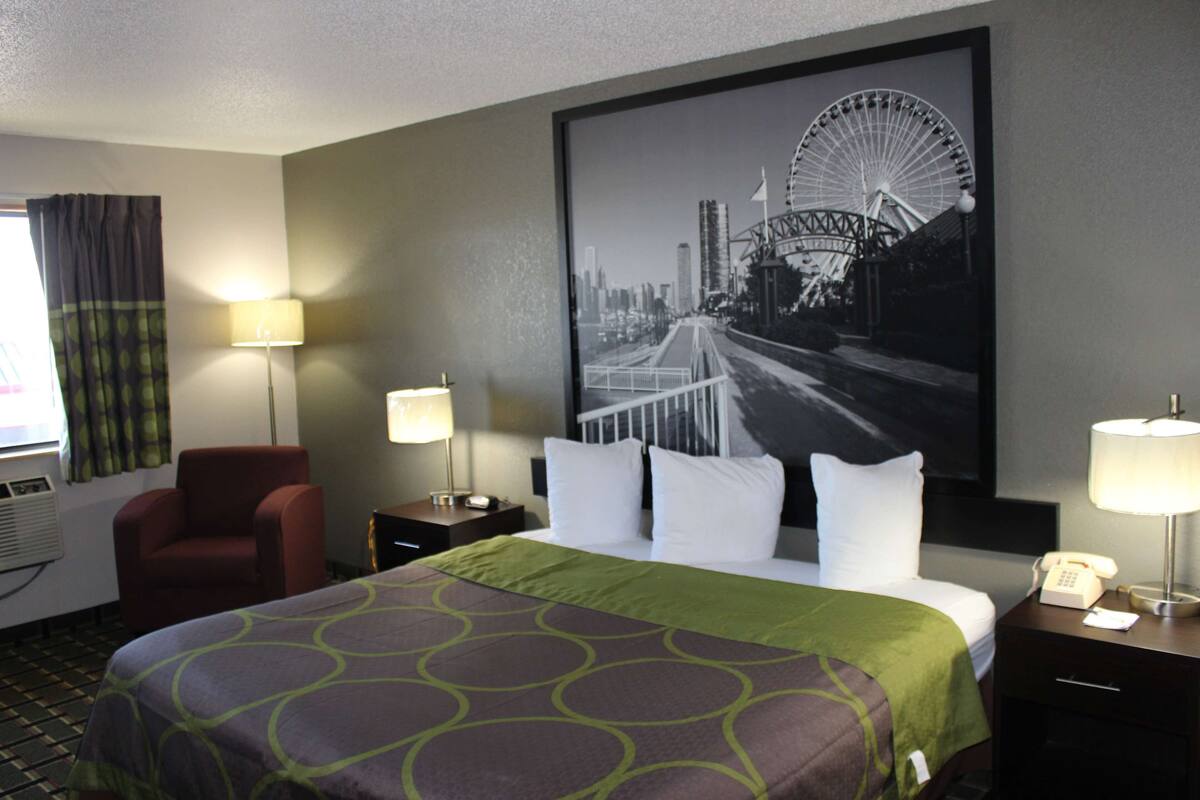 A hotel room with a large bed, two bedside tables with lamps, a red armchair, a wall-mounted air conditioner, and a large cityscape poster on the wall.