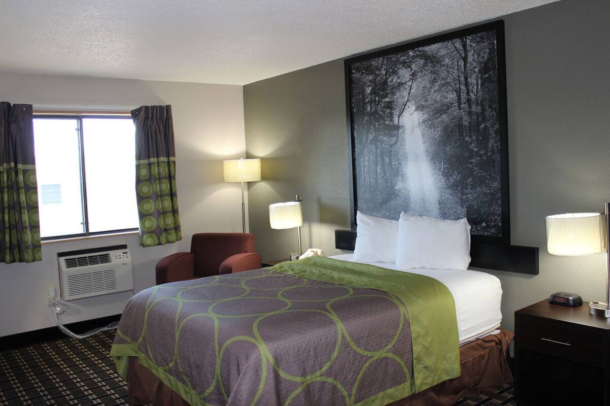 A hotel room with a bed, two bedside lamps, a chair, an air conditioning unit, and a large painting on the wall.