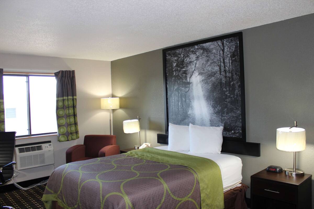 A hotel room with a bed, two bedside lamps, a window with curtains, a wall-mounted air conditioner, and a large framed picture of a forest scene.