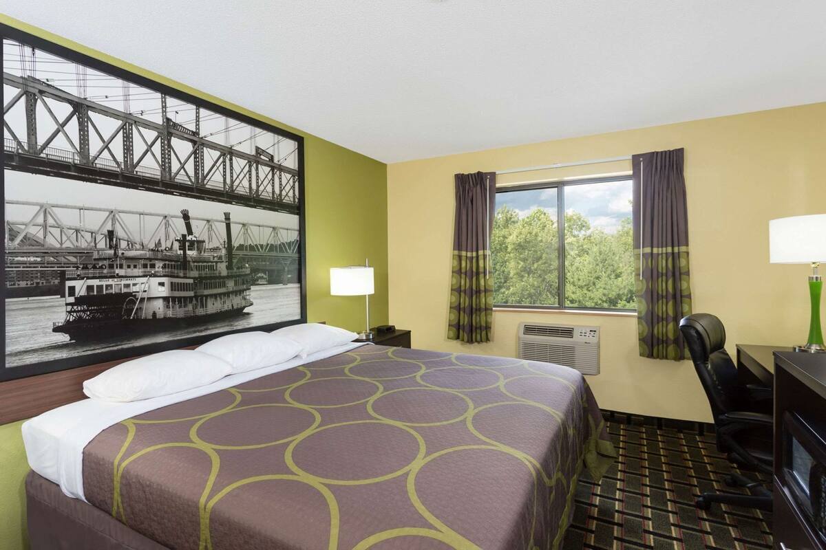 Habitación de hotel con cama, escritorio, silla, ventana con cortinas, unidad de aire acondicionado y un cuadro grande con un puente y un barco.