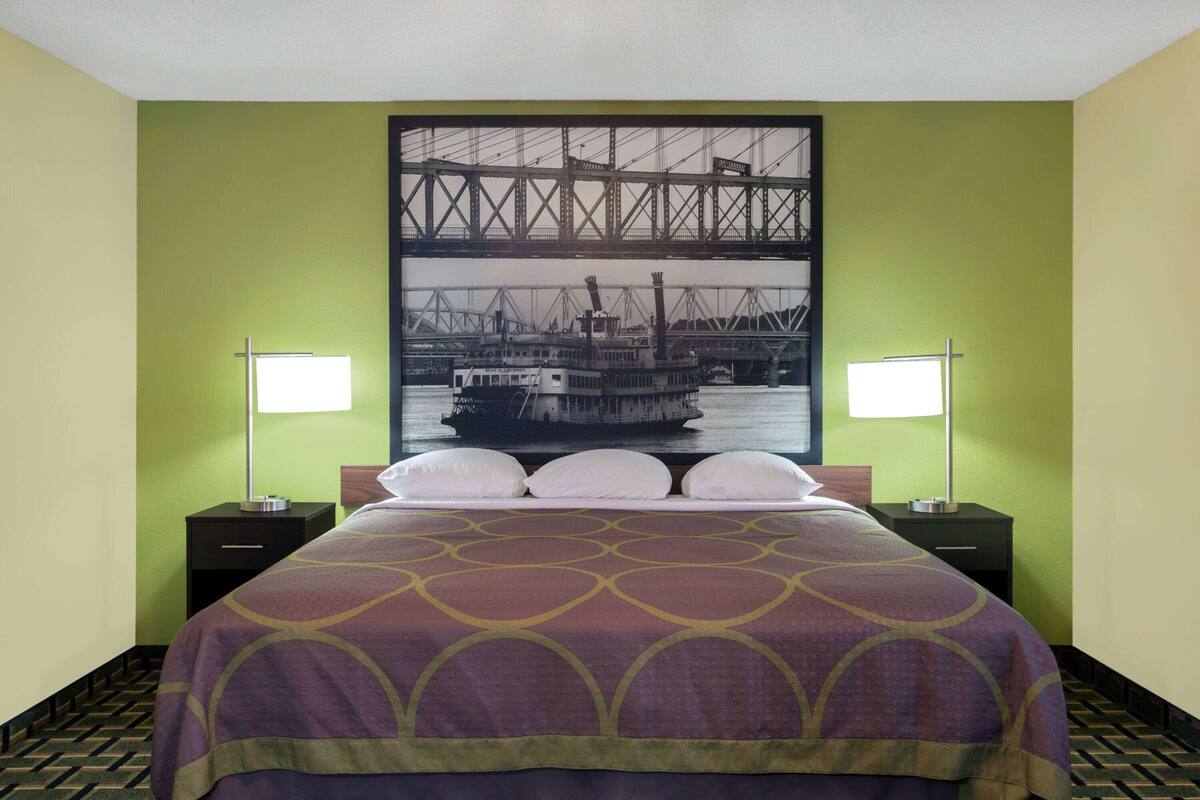 Une chambre d’hôtel avec un lit, deux tables de chevet et un grand tableau encadré représentant un pont et un bateau, accroché au mur.