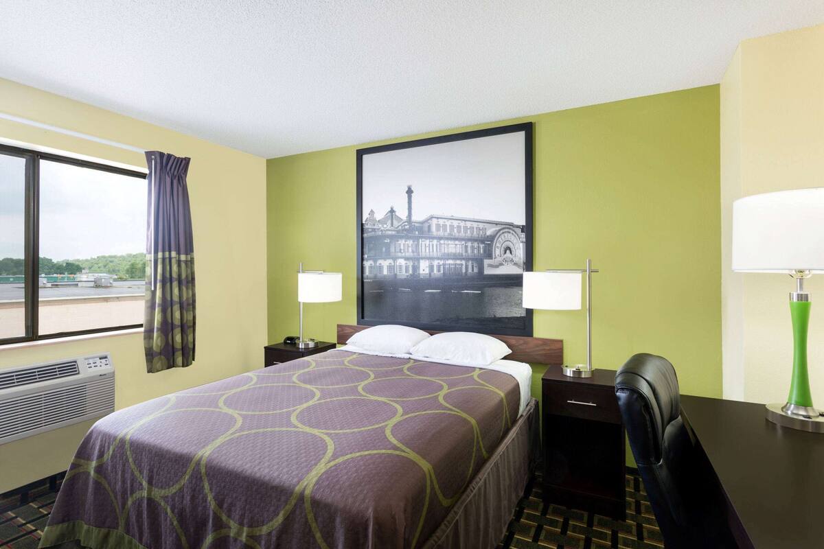 Une chambre d’hôtel comprenant un lit, un bureau, une chaise, une fenêtre avec des rideaux, un climatiseur fixé au mur et un tableau encadré accroché au mur.