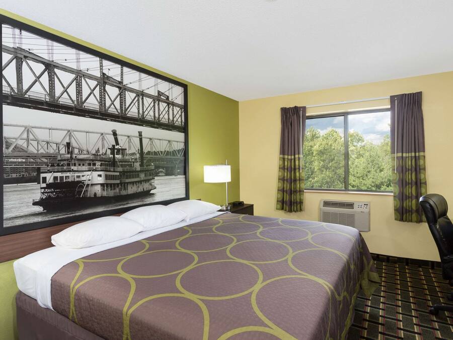 A hotel room with a bed, a desk, a chair, a window with curtains, a lamp, and a wall-mounted air conditioner.