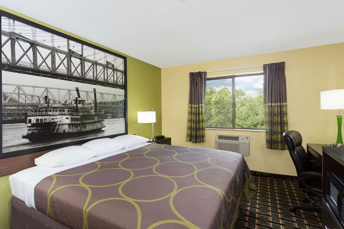 Une chambre d’hôtel comprenant un lit, un bureau, une chaise, une fenêtre avec des rideaux, un appareil de climatisation et un grand tableau représentant un pont et un bateau.