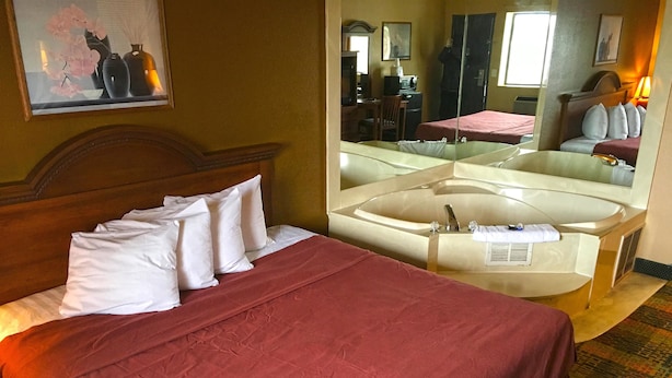 A hotel room with a bed, a large mirror, a desk, and a bathroom with a bathtub. - Americas Best Value Inn Livonia Detroit (Livonia)