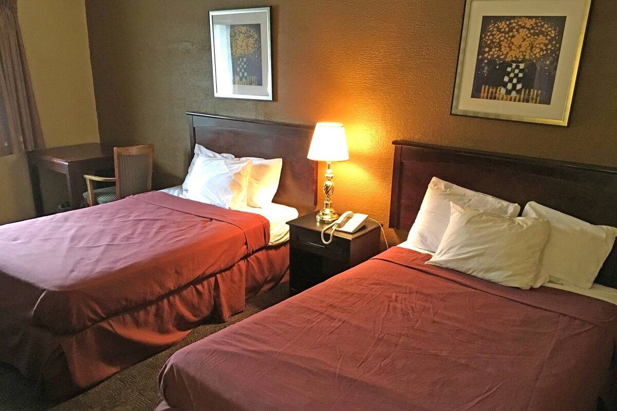 A hotel room with two beds, each with a red bedspread and white pillows. There is a nightstand with a lamp and a phone between the beds. The room has a framed picture on the wall above the beds.