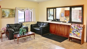 A living room with two black leather sofas, a wooden coffee table, a potted plant, a window with curtains, a bulletin board with various notices, and a reception desk with a mirror. - Americas Best Value Inn Livonia Detroit (Livonia)
