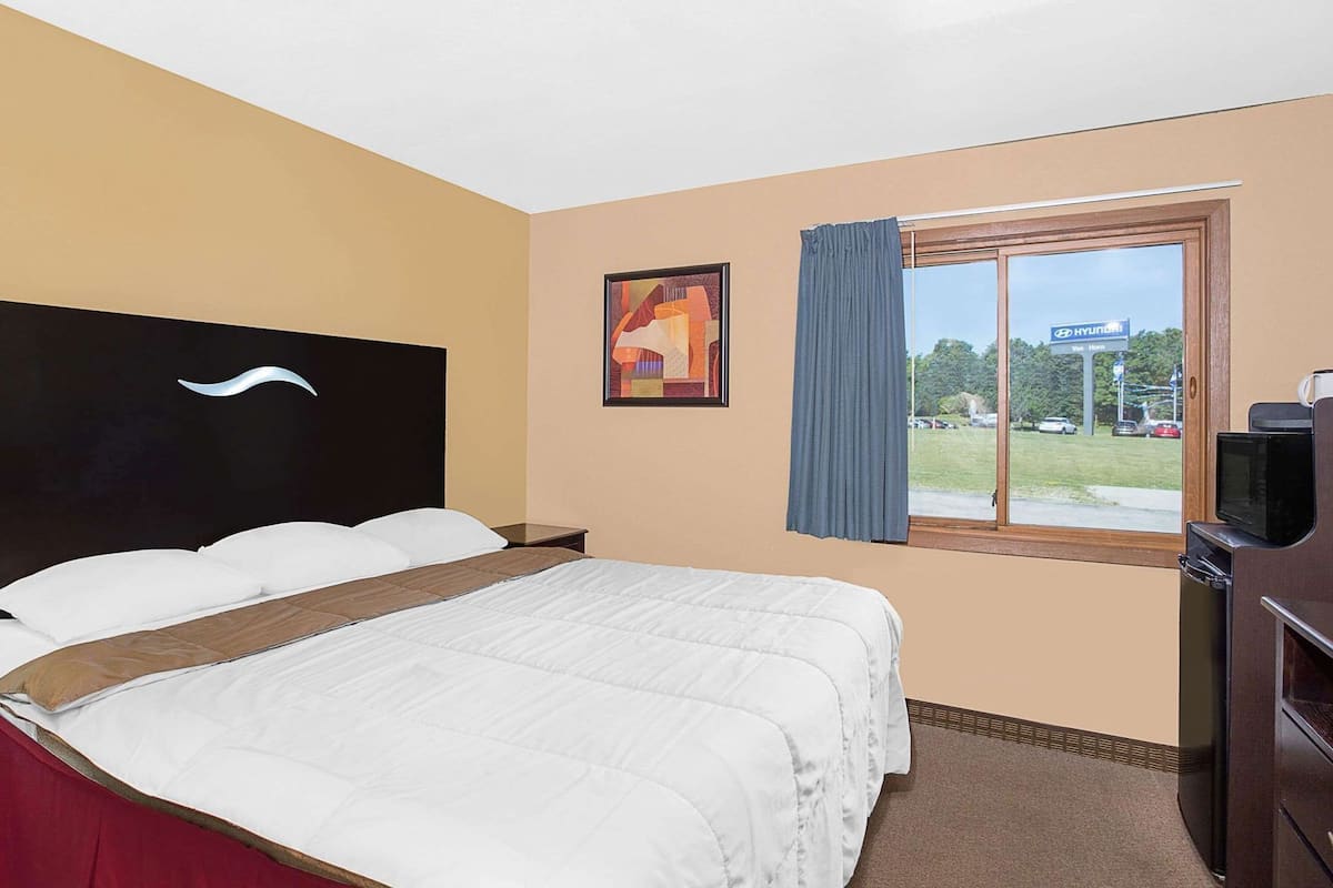 A hotel room with a bed, a television, a window with a view of a parking lot, and a painting on the wall.