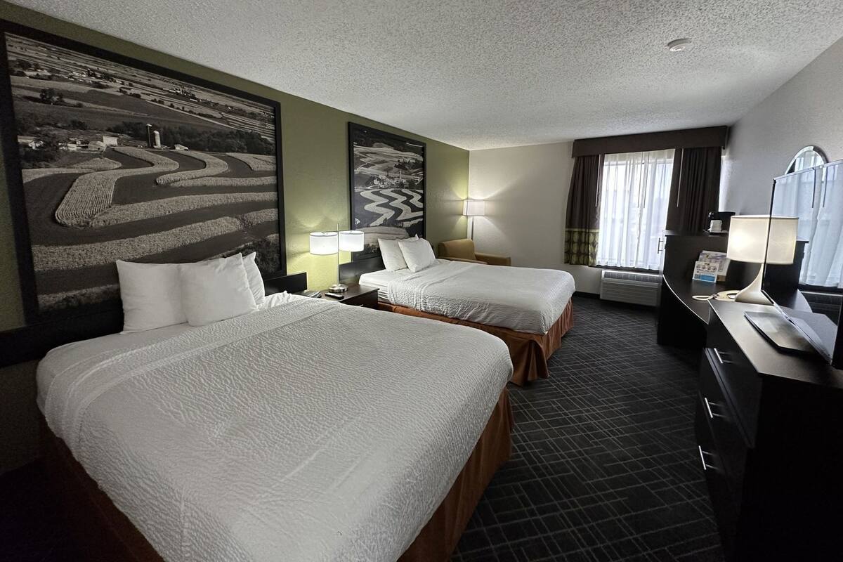 A hotel room with two beds, a desk, a chair, two lamps, a large painting on the wall, and a window with curtains.