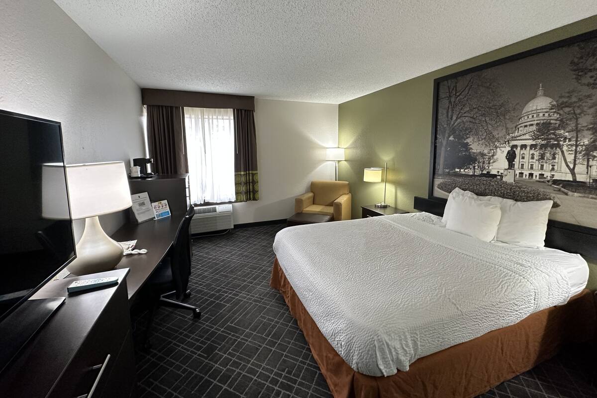 A hotel room with a bed, desk, chair, and a large wall painting of a building.