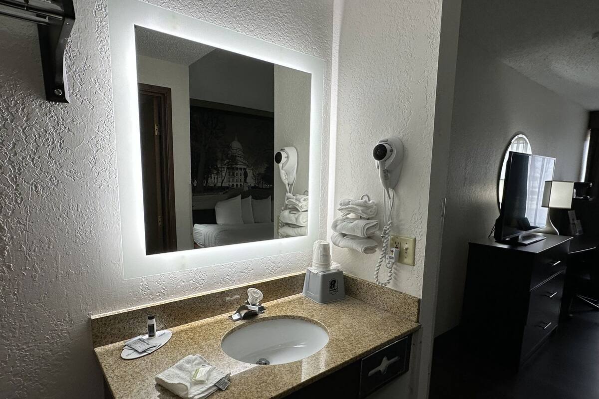 A hotel bathroom with a large mirror, a sink, a hairdryer, and a desk with a computer and lamp.