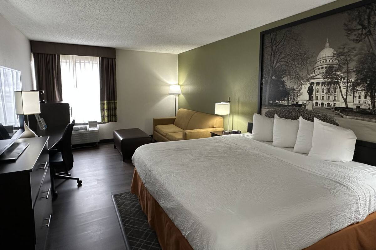A hotel room with a large bed, a desk with a chair, a sofa, and a painting of a building on the wall.