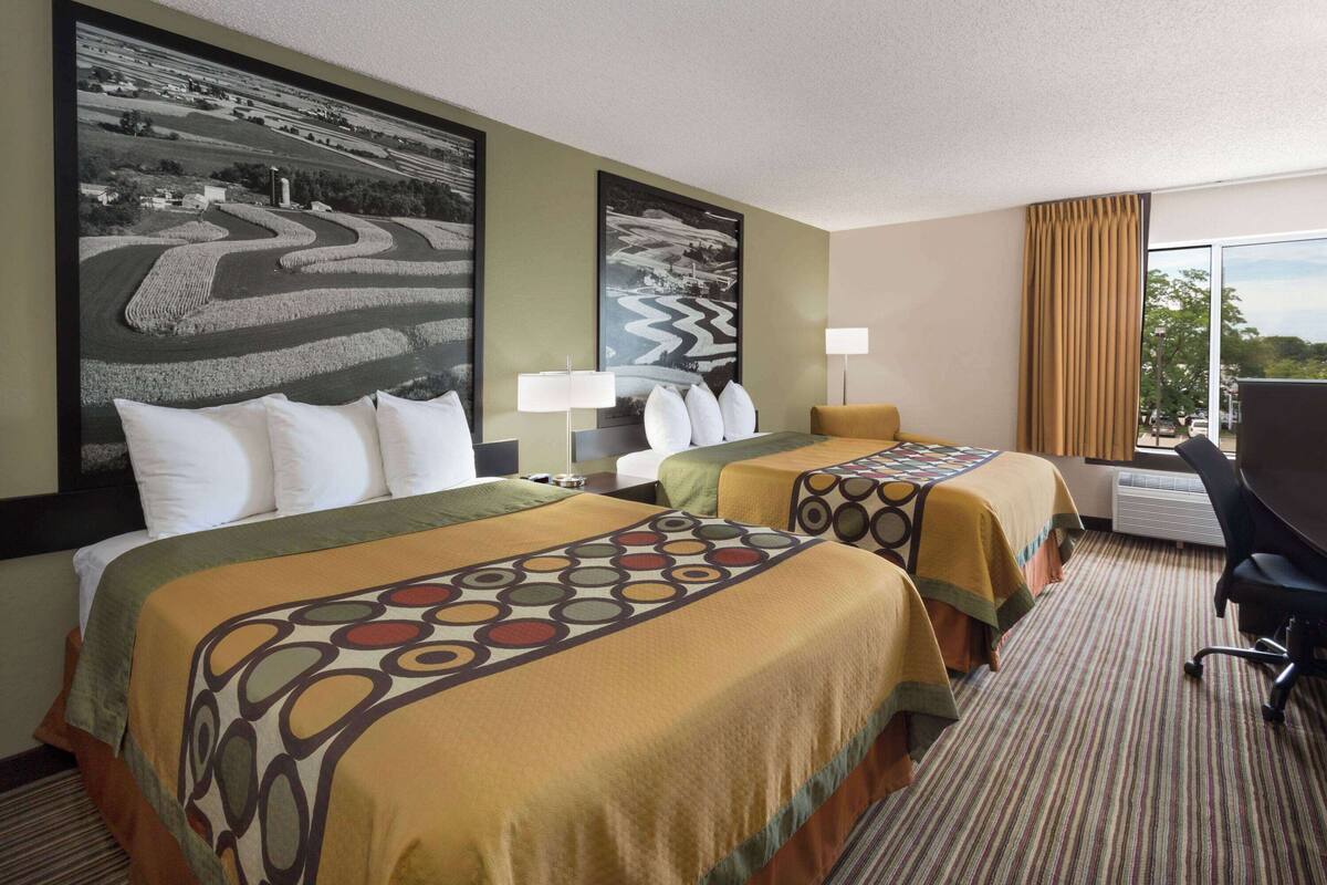 A hotel room with two beds, large landscape paintings, a desk with a chair, and a large window with curtains.