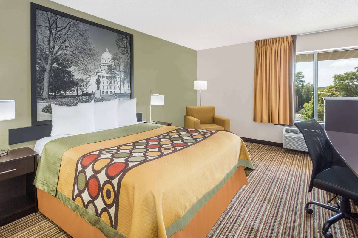 A hotel room with a large bed, a desk, a chair, a lamp, and a large window with curtains.