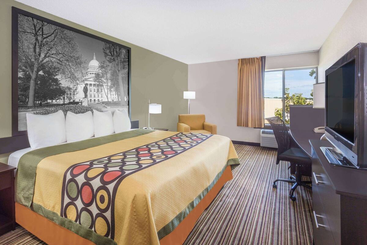 A hotel room with a large bed, a desk with a computer, a television, and a painting of a building on the wall.