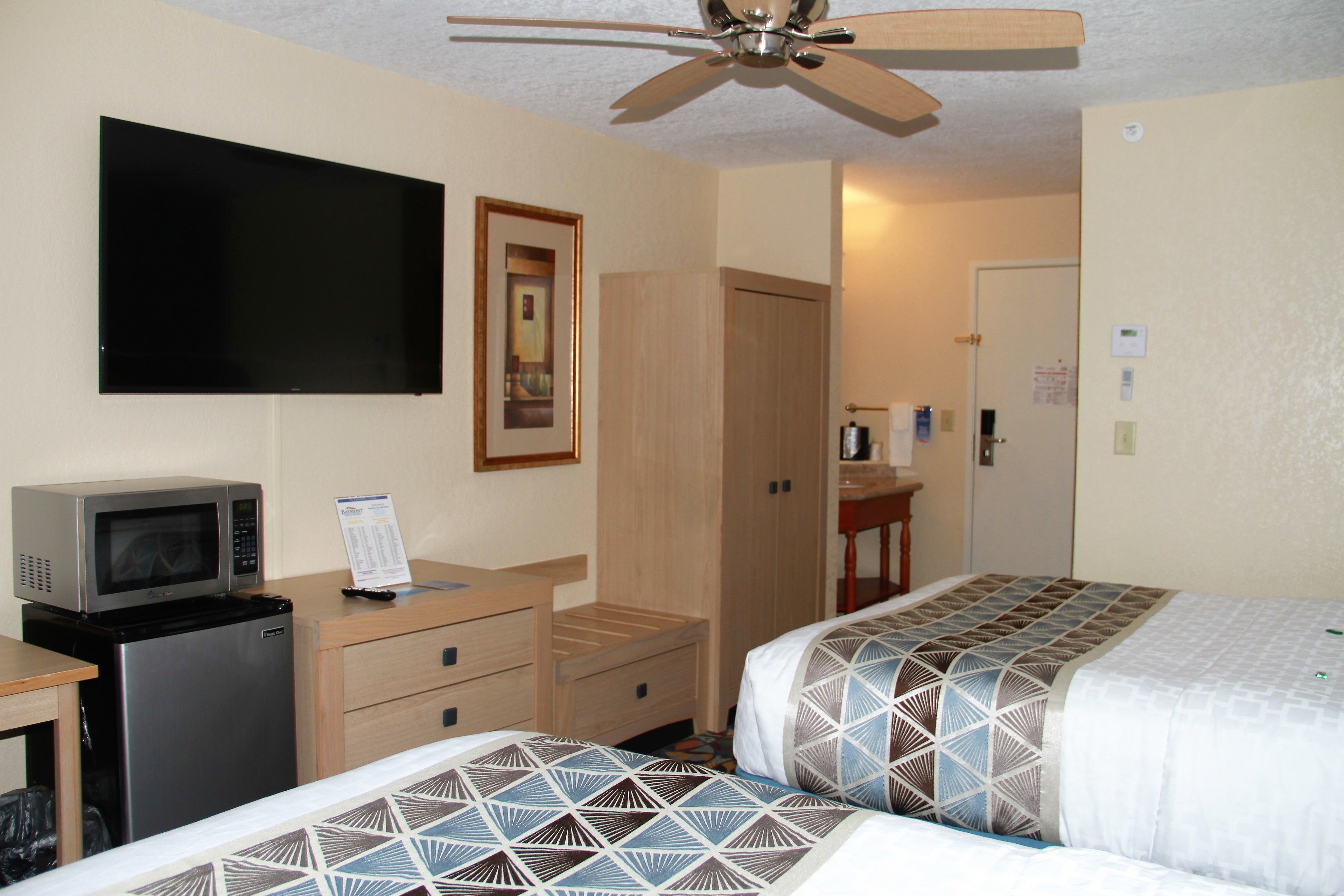 Une chambre d’hôtel équipée d’un lit, d’un bureau avec un micro-ondes, d’un ventilateur de plafond et d’un téléviseur fixé au mur.