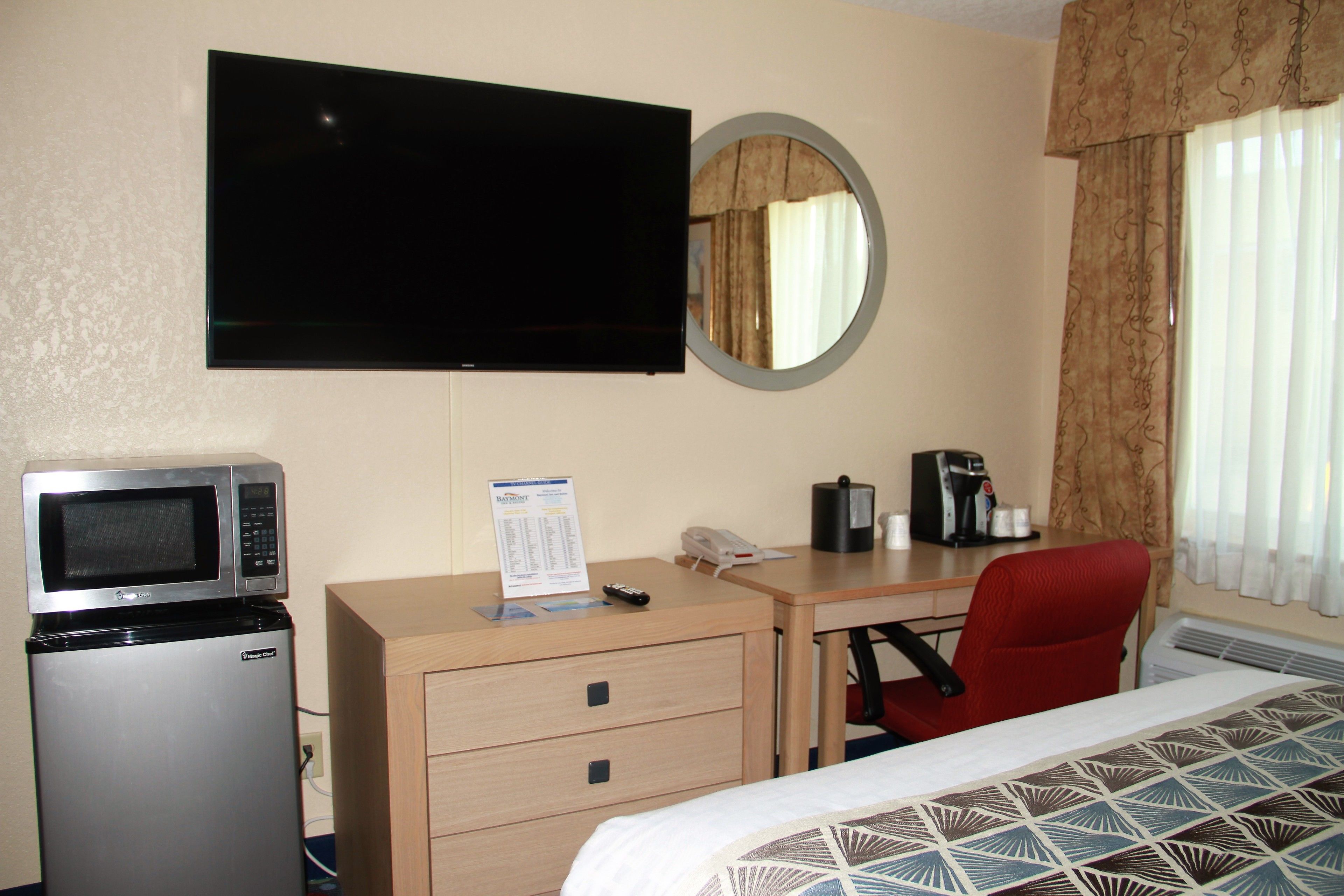 Une chambre d’hôtel comprenant un lit, un bureau avec une chaise, un four à micro-ondes, un réfrigérateur, une télévision, un miroir et une fenêtre avec des rideaux.