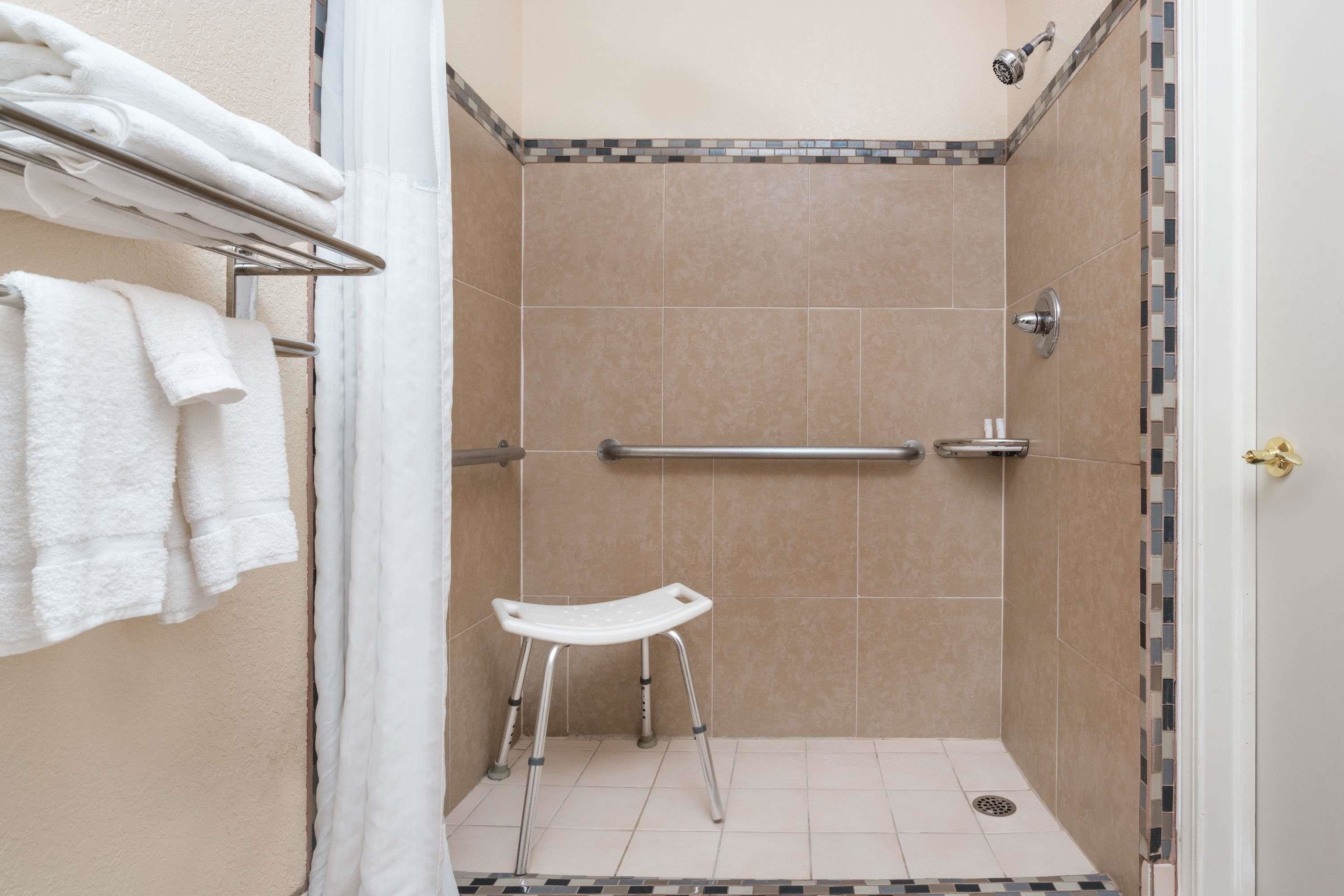 Un área de ducha con azulejos color beige, un asiento de ducha blanco, un perchero con toallas dobladas y una puerta con pomo dorado.