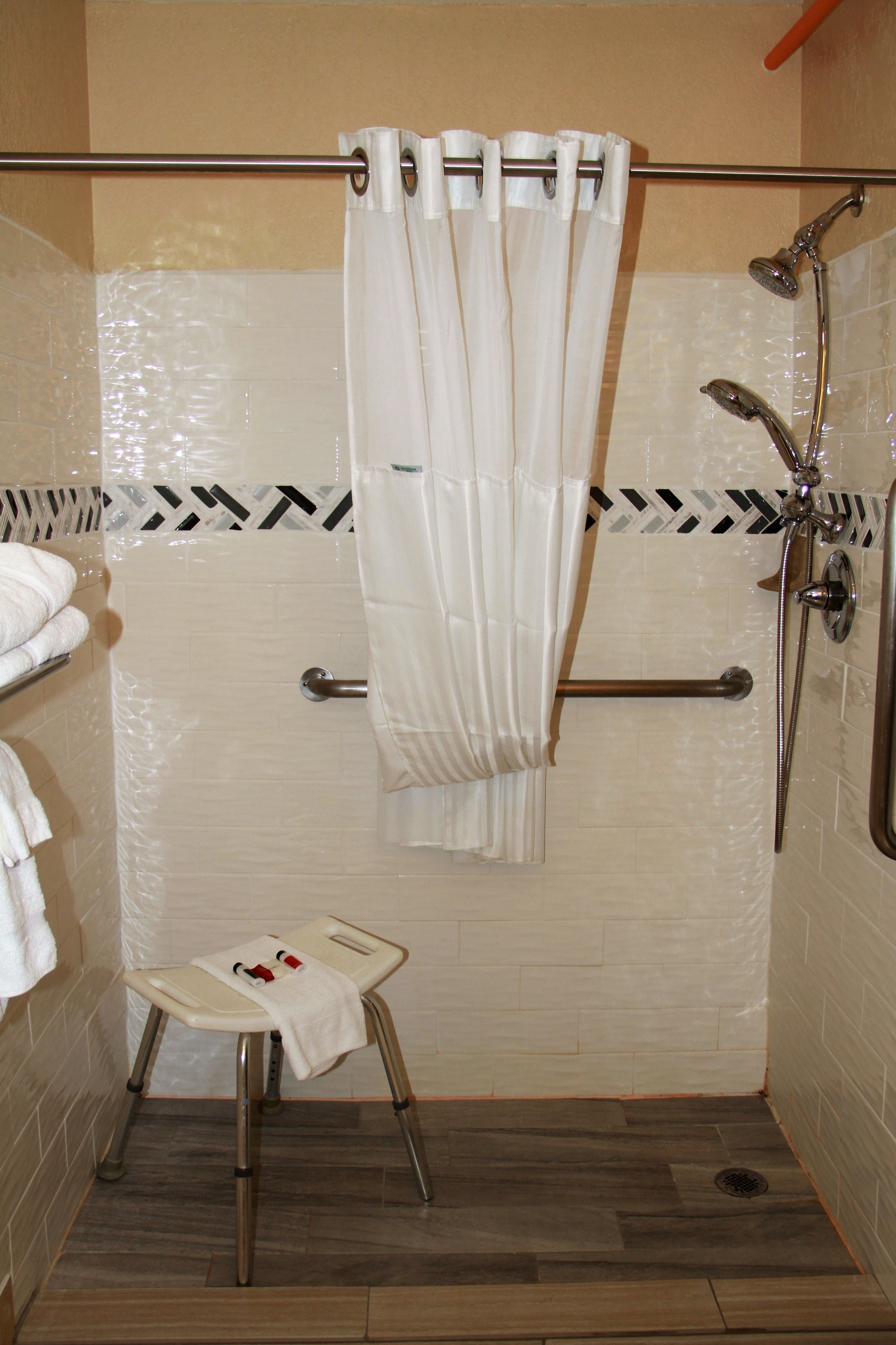 A shower area with a white shower curtain, a handheld showerhead, a grab bar, and a small stool with a towel.