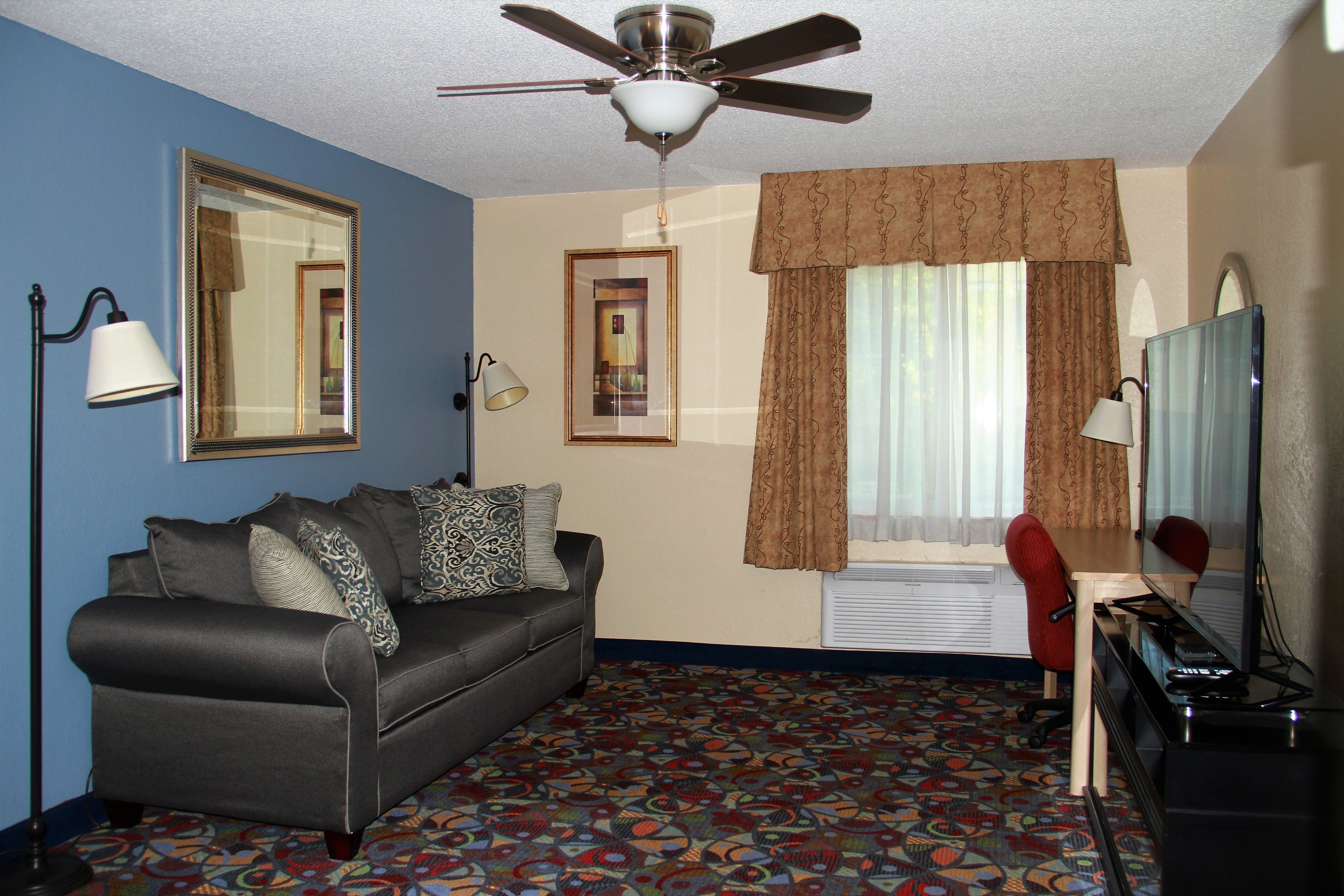 A hotel room with a ceiling fan, a sofa, a desk with a chair, a large mirror, and patterned carpet.