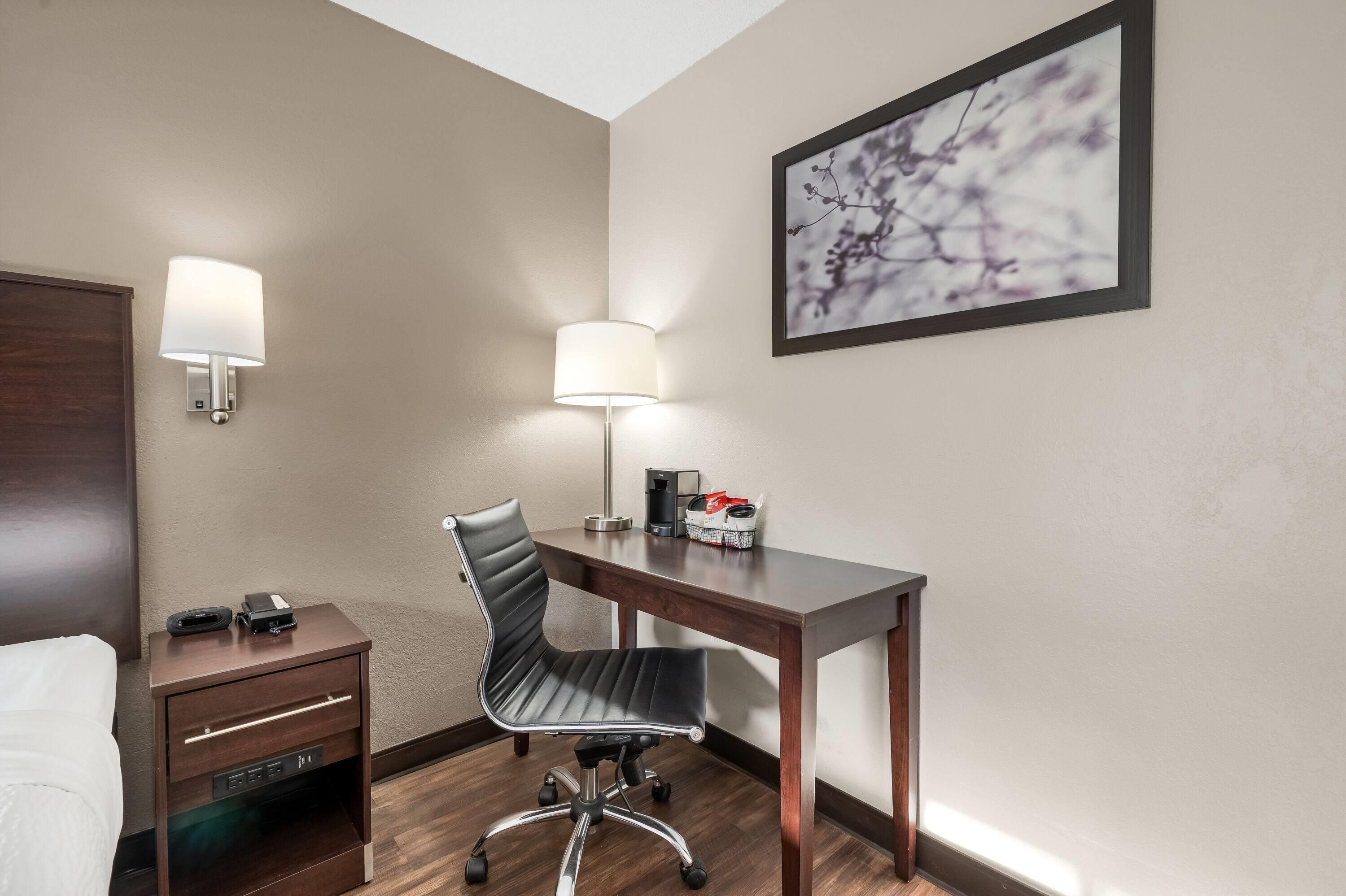 Une chambre d’hôtel avec un lit, un bureau, une chaise, une table de chevet, une lampe et un tableau encadré au mur.