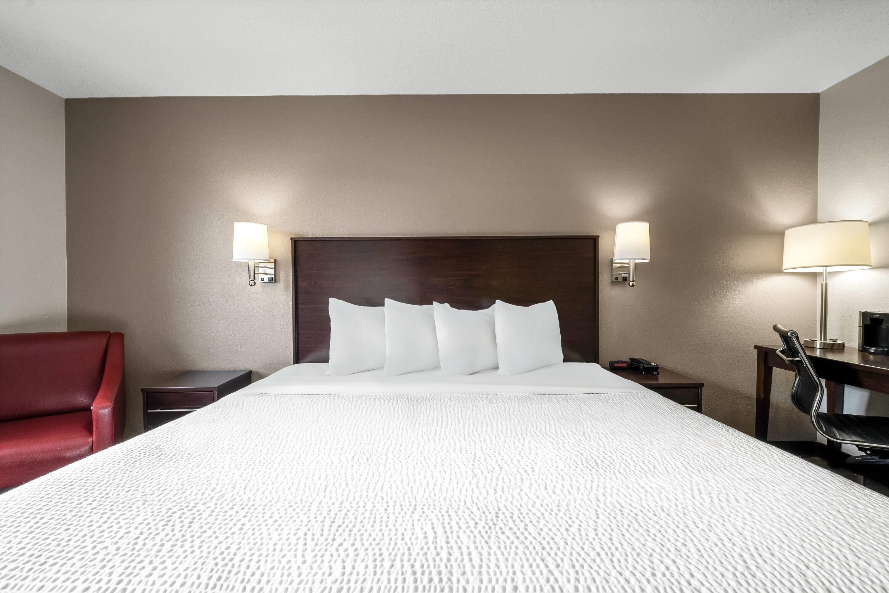 A hotel room with a large bed, two bedside tables, a red chair, and a desk with a lamp.