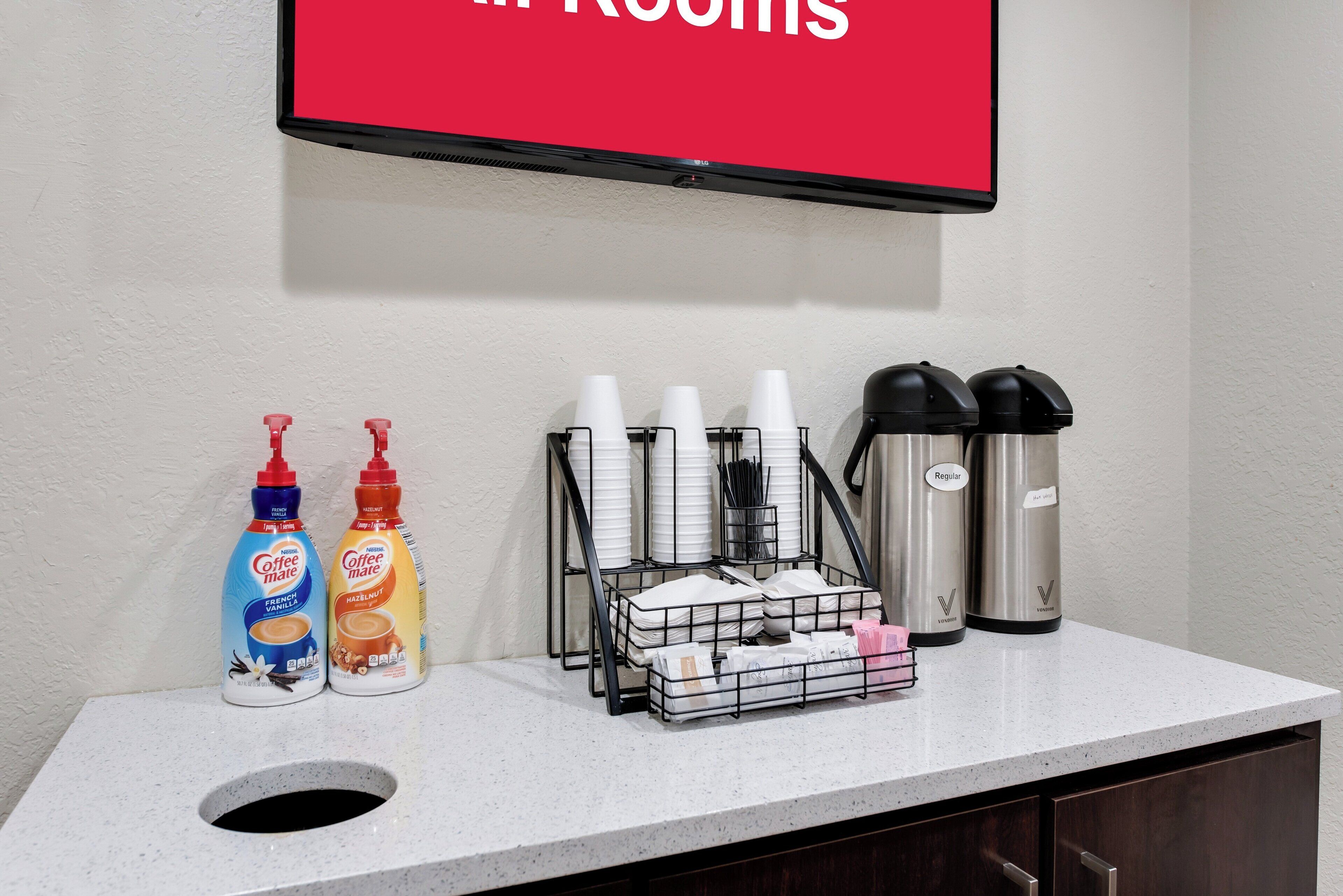 A hotel reception area with a flat-screen TV displaying "ROOMS" above a counter. On the counter, there are two bottles of Coffee Mate, a coffee machine, a tray with cups and napkins, and two thermos flasks.