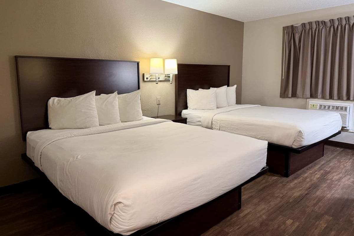 A hotel room with two beds, a nightstand, a lamp, and a window with curtains.