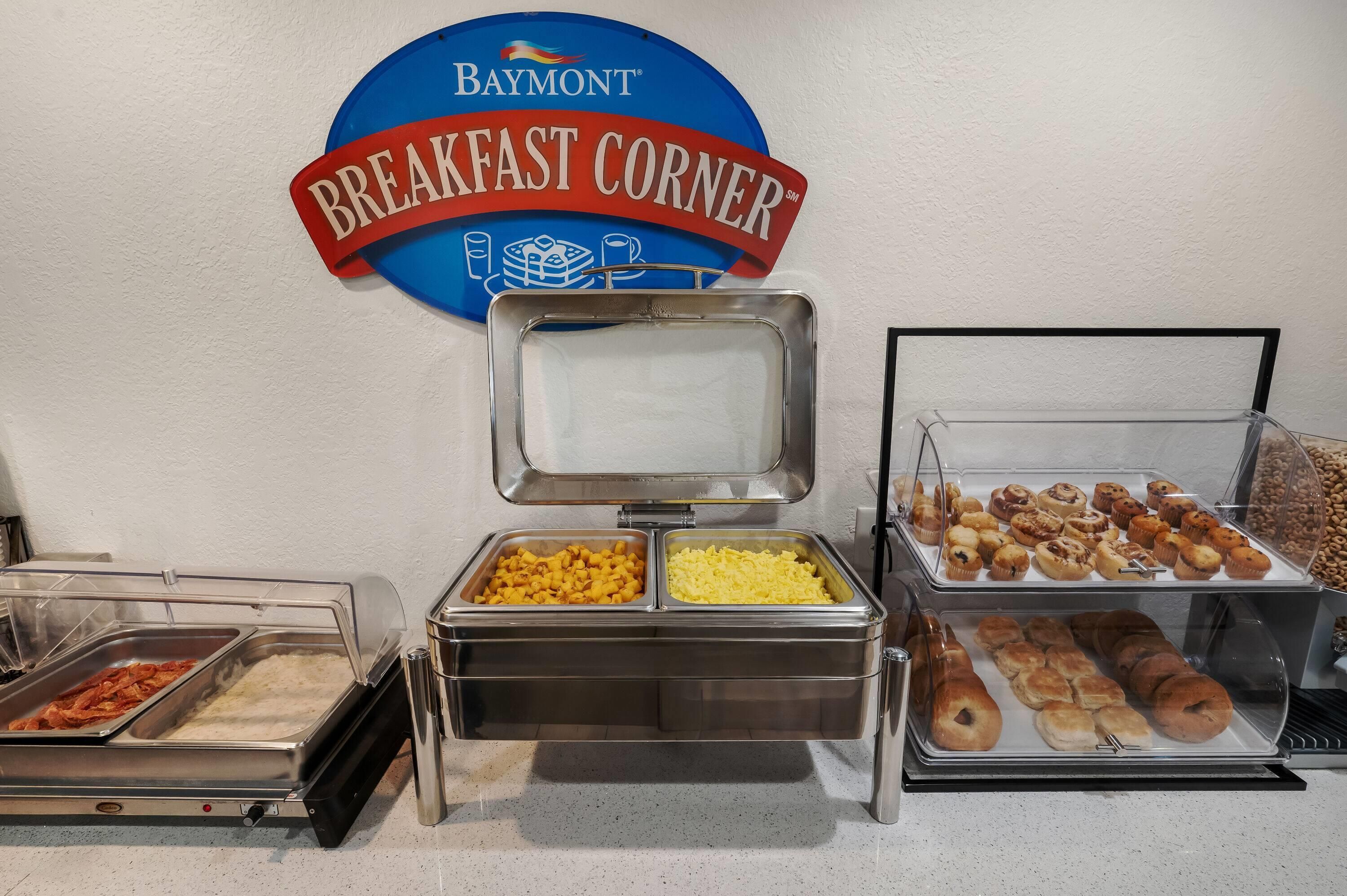 Zona de desayuno tipo buffet con un letrero que dice 'Baymont Breakfast Corner', que ofrece diversas opciones de comida como bollería y frutas.