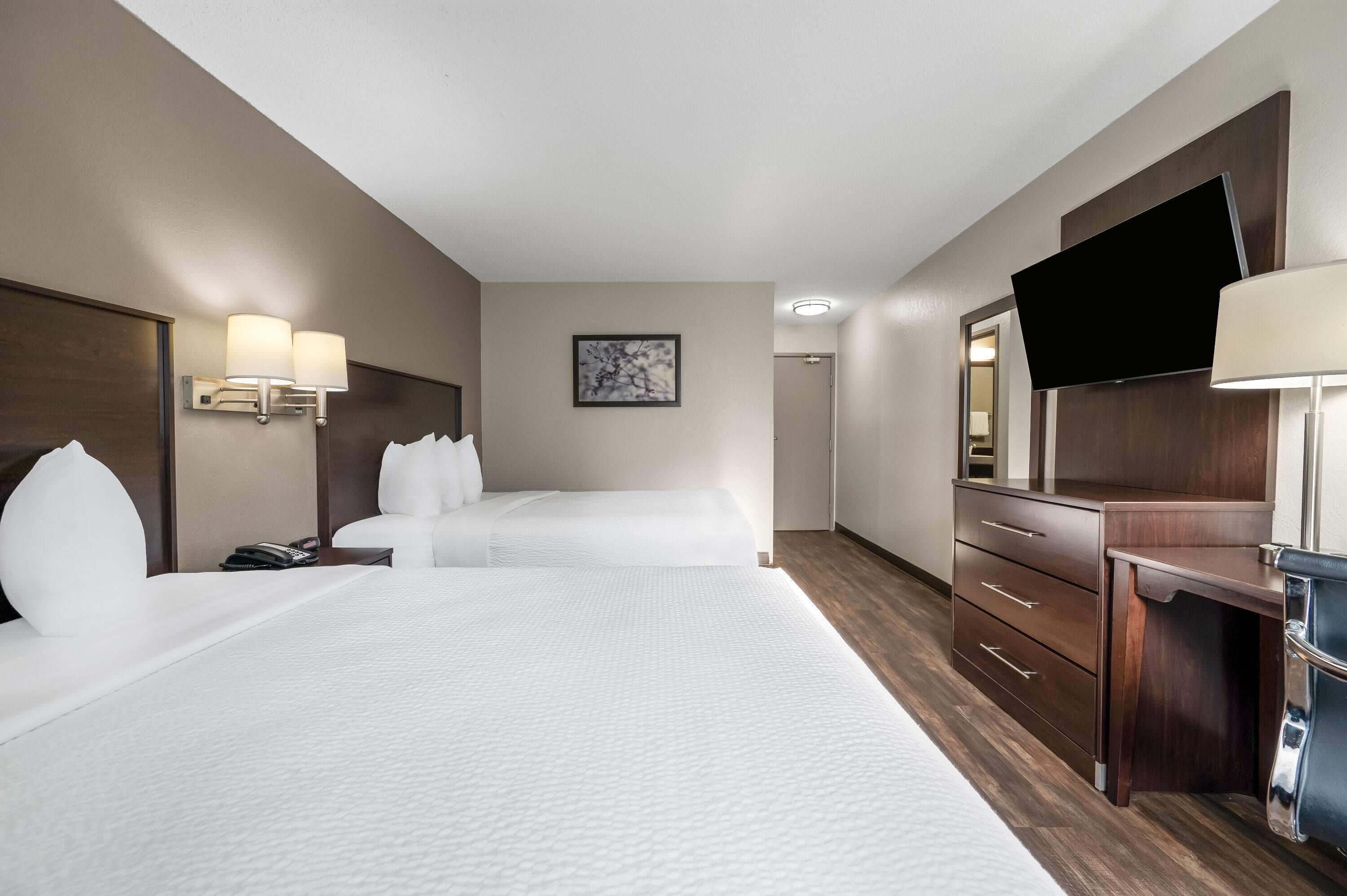 A hotel room with two beds, a desk, a television, and a wall-mounted lamp.