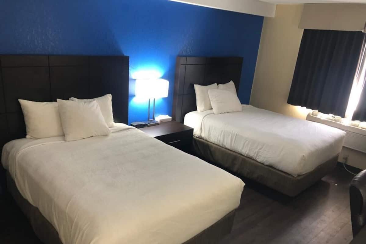 A hotel room with two beds, each with white linens and headboards. There is a bedside table with a lamp on one side and a window with curtains on the other.